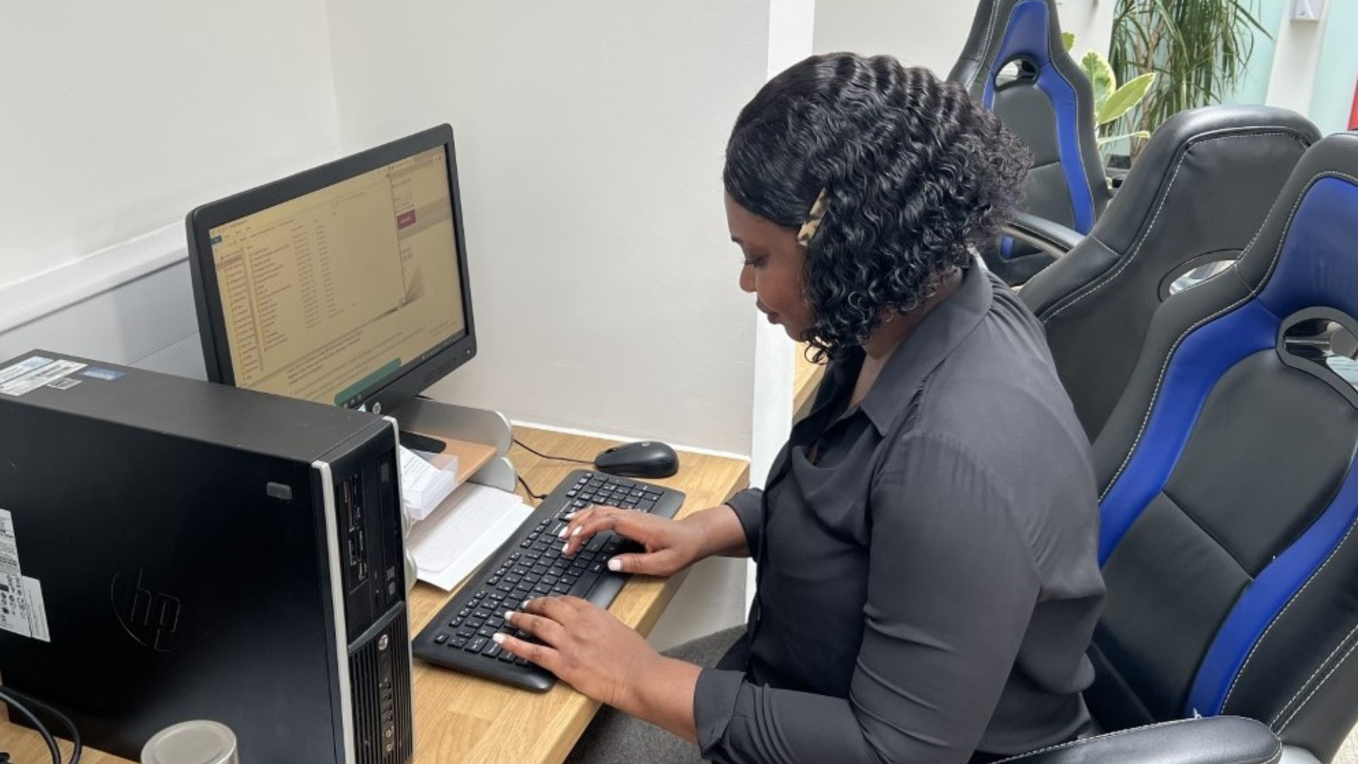A woman with curly black hair sitting at a desk working on a computer in an office. She is wearing a dark gray shirt, using a keyboard and mouse, with a monitor, a desktop computer tower, and office chairs nearby. There is also a plant in the background.