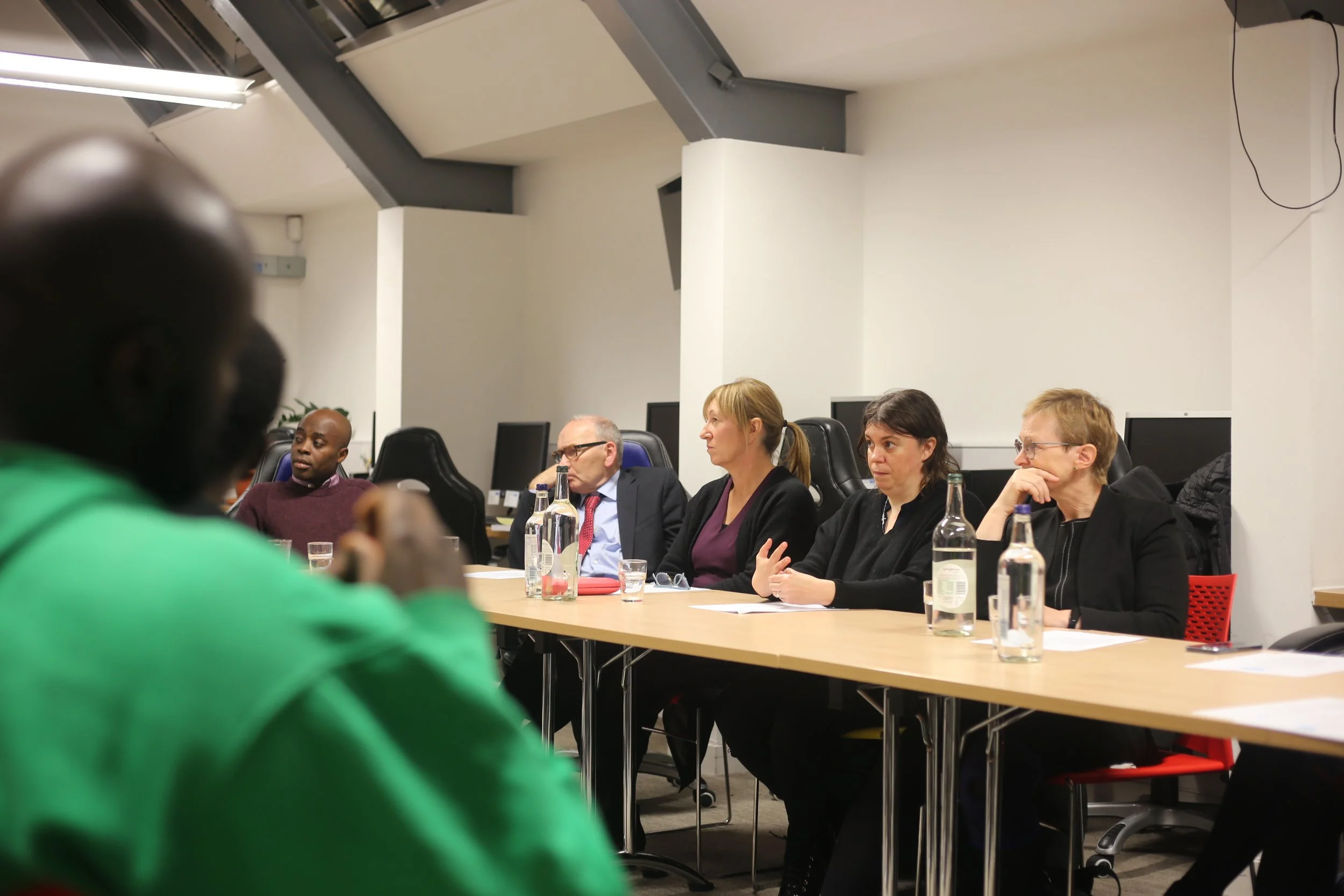 A group of five people seated at a conference table in a meeting room. Four women and one man, with three women engaging and speaking, while the man appears to be listening. Bottles of water and glasses are on the table.
