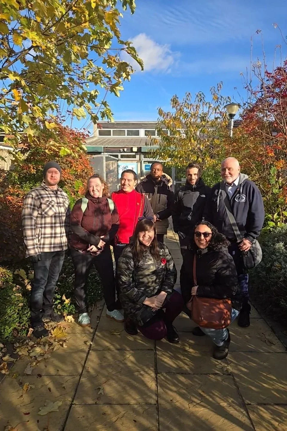 A group of eight people standing outdoors on a sidewalk surrounded by fall foliage, smiling for a photo on a sunny day.