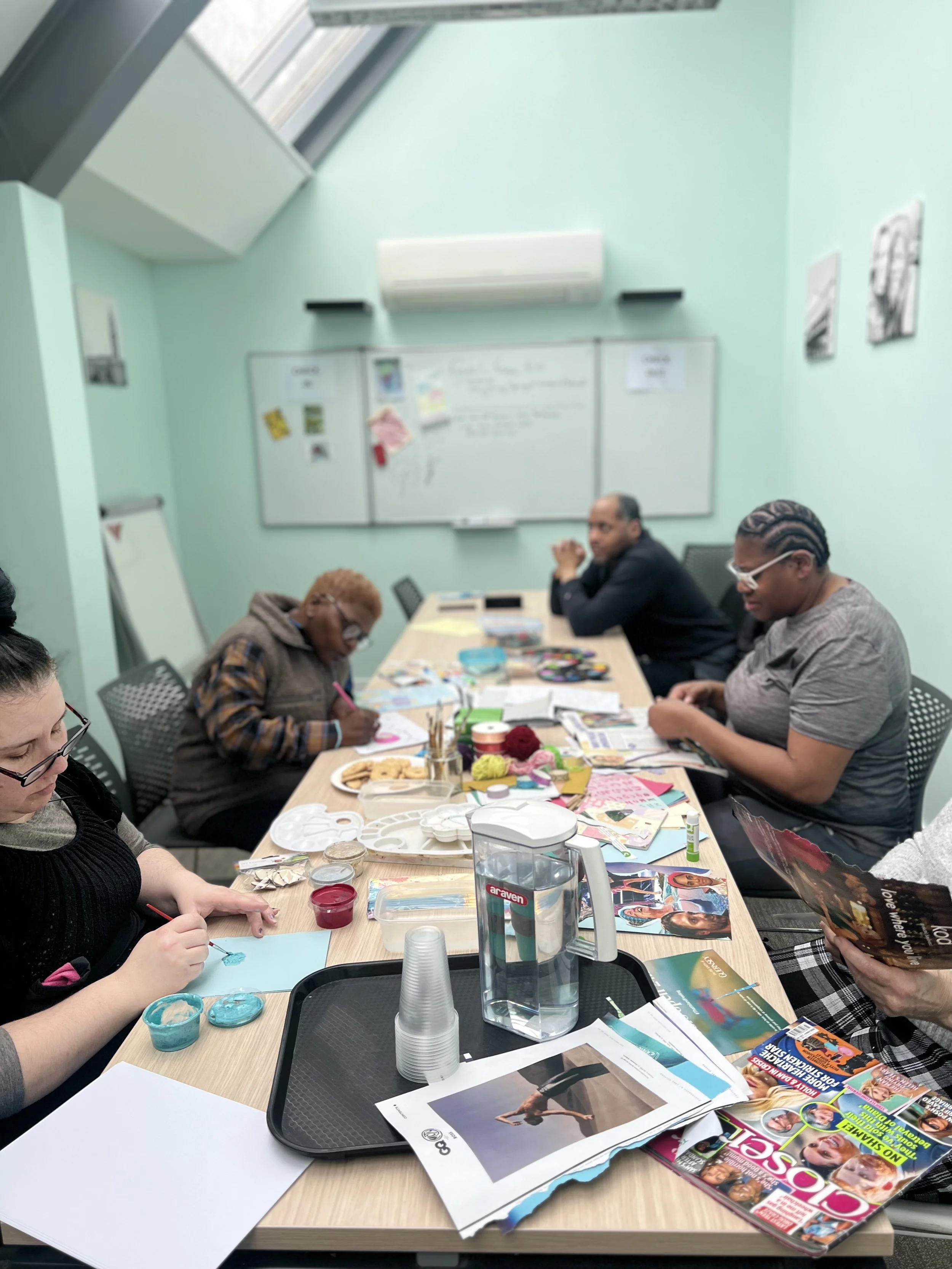People participating in an arts and crafts workshop around a table with art supplies, magazines, and snacks in a room with mint green walls and a whiteboard.