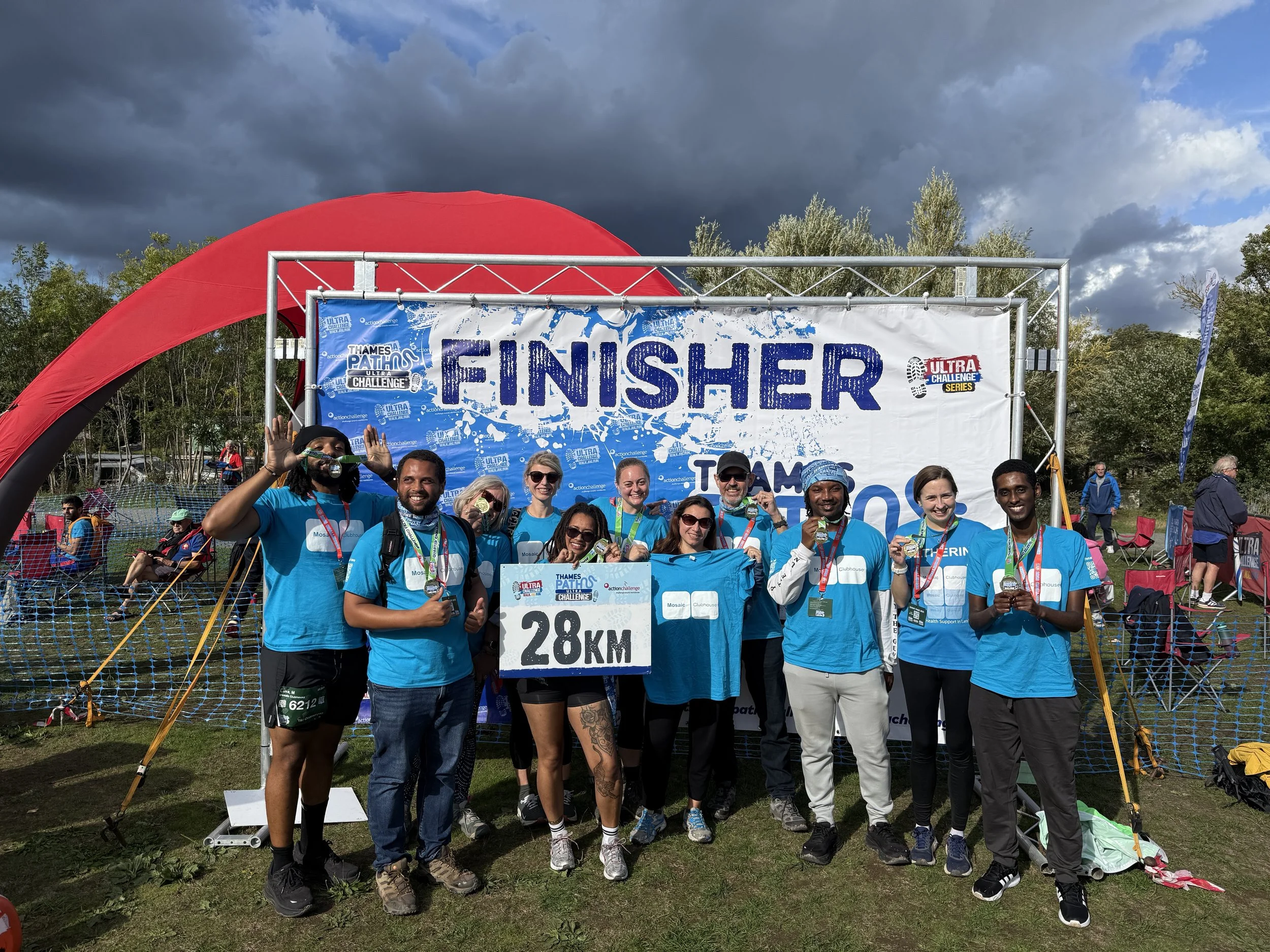 Group of people in blue shirts celebrating at the finish line of a 28 km race, with a large blue and white 'FINISHER' banner and a partially cloudy sky in the background.