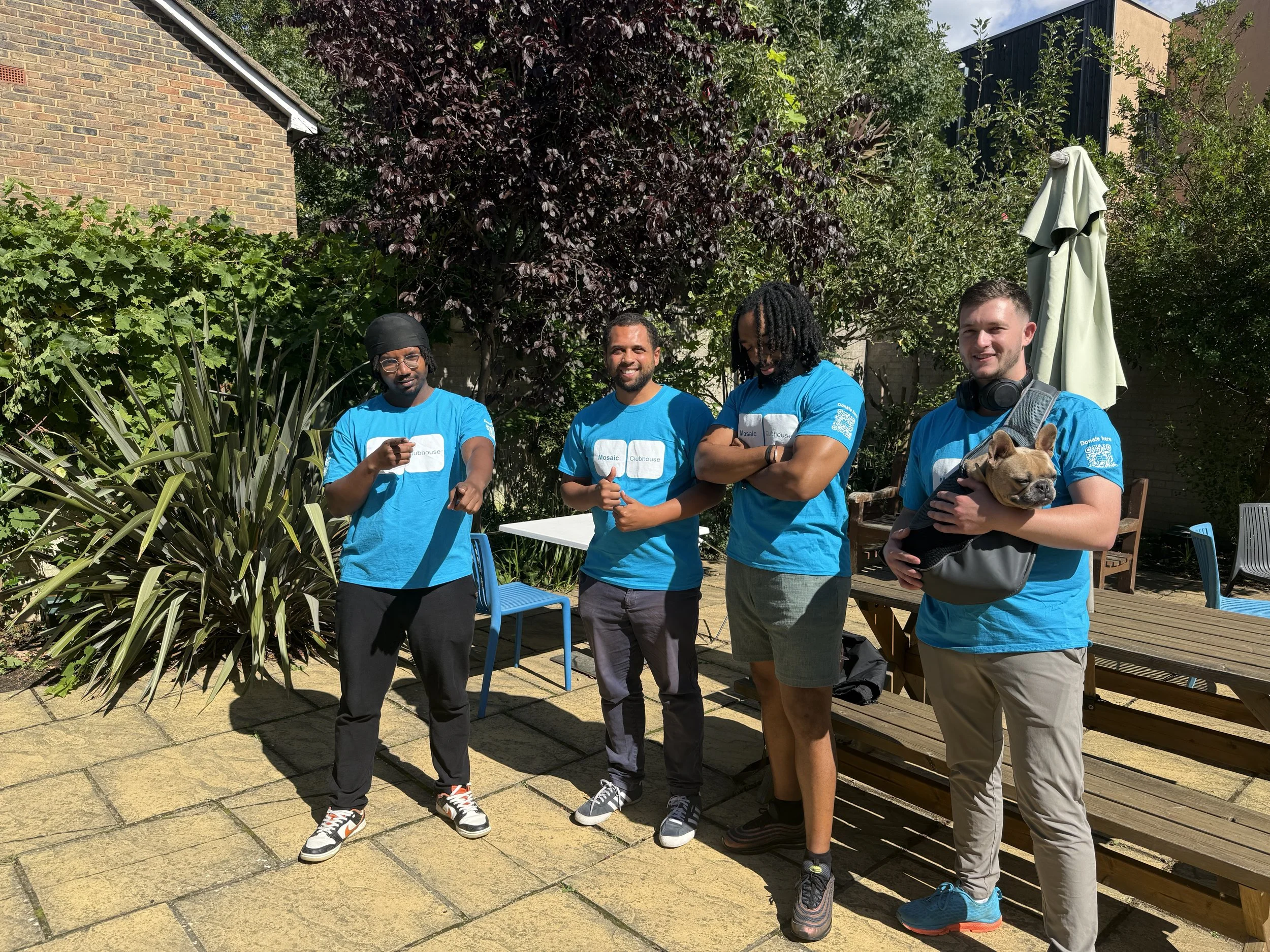 Four men in matching blue t-shirts standing outdoors in a garden or backyard area, some smiling and holding a dog, with trees, plants, and a brick and black building in the background.