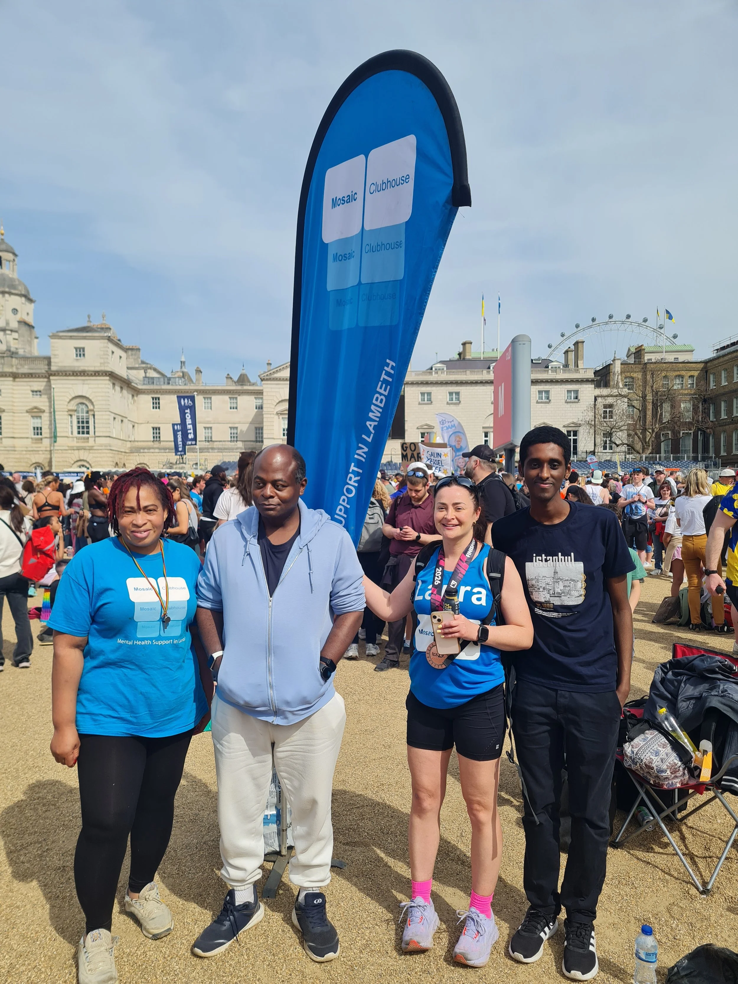 A diverse group of four people standing outdoors at a crowded event, with historical buildings and a Ferris wheel in the background. Some are wearing shirts supporting mental health and standing in front of a blue flag.