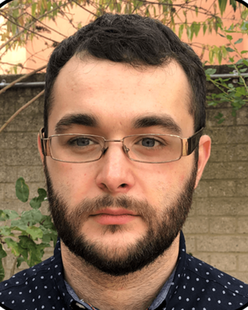 Close-up of a young man with dark hair, glasses, and a beard, standing outdoors with a brick wall and green foliage in the background.