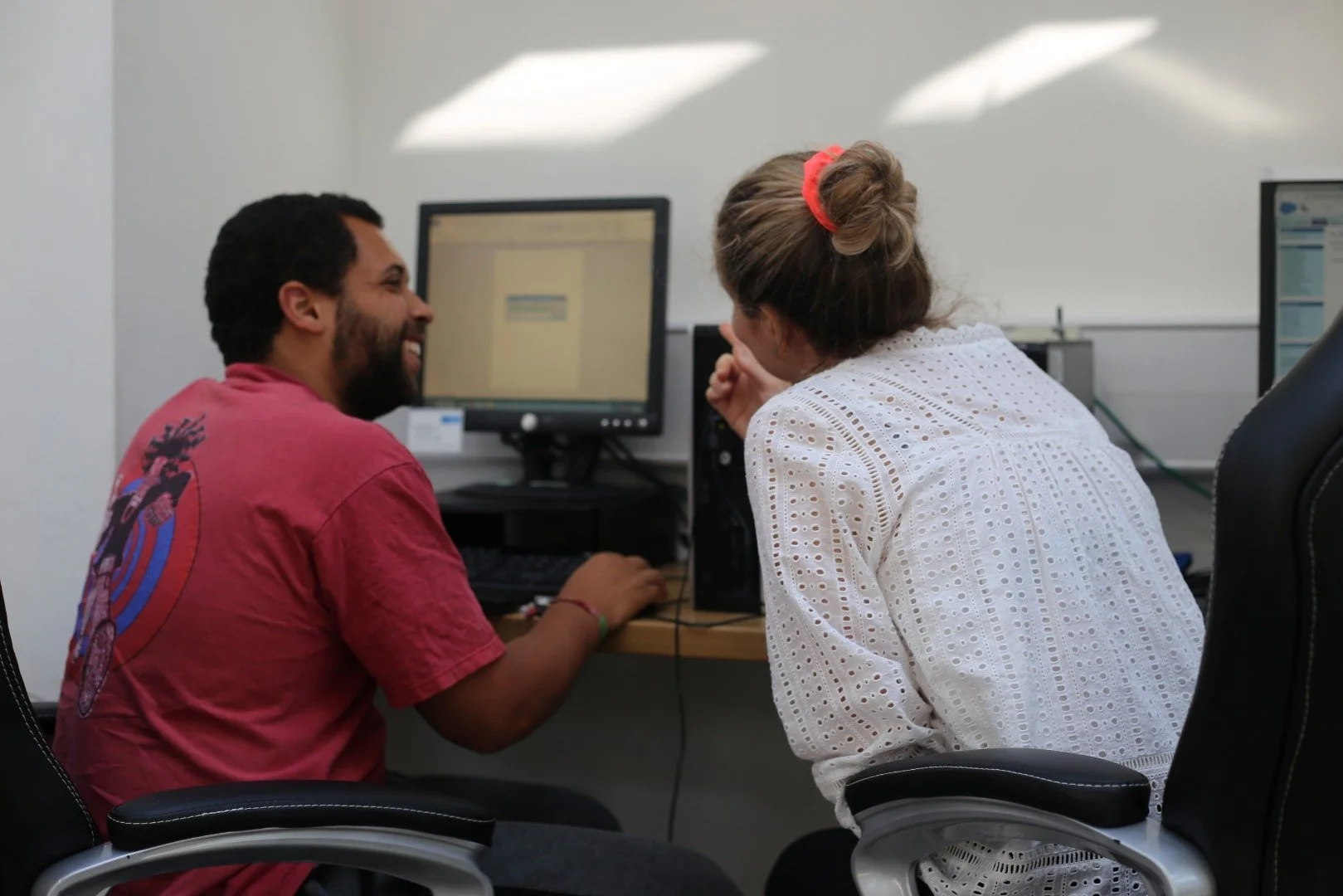 A man and woman are sitting at a desk in an office, laughing and talking. The man is using a computer, and they seem to be engaged in a lively conversation.