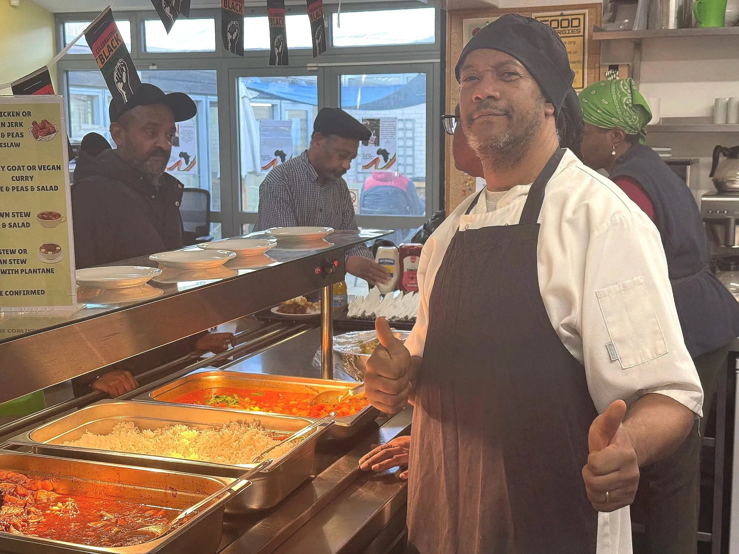 Chef in white uniform and black apron giving a thumbs-up in a cafeteria-style setting with food trays and other staff in the background.