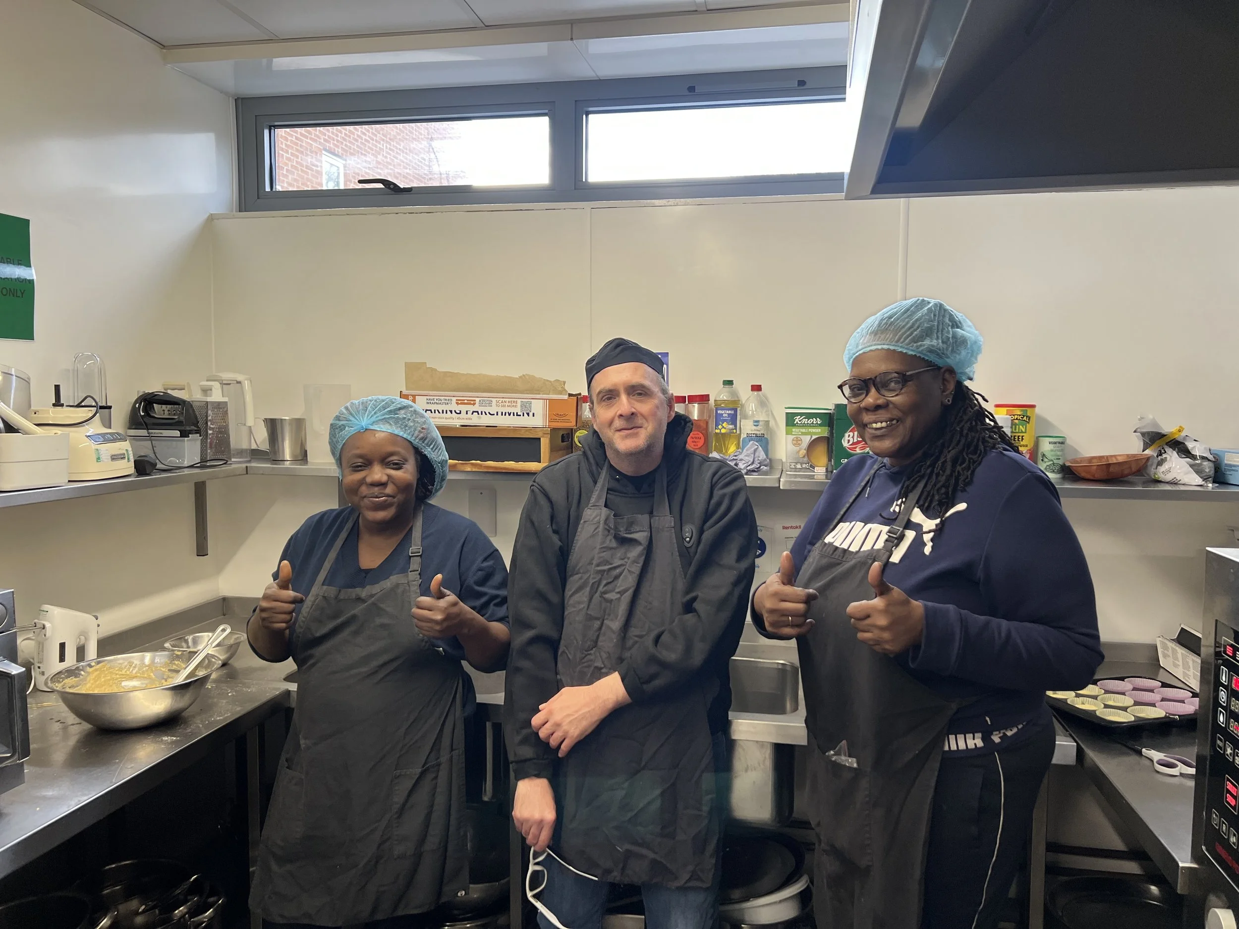 Three people in a kitchen posing together, giving thumbs up. They are wearing hairnets and aprons, standing behind a kitchen counter with baking ingredients and tools, including a bowl of batter, cupcake liners, and kitchen appliances.