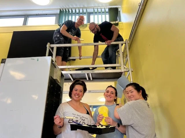 Three women standing on the ground holding a cake and a yellow plush toy, and two men standing on a platform above them working on a structure in a yellow room with windows.
