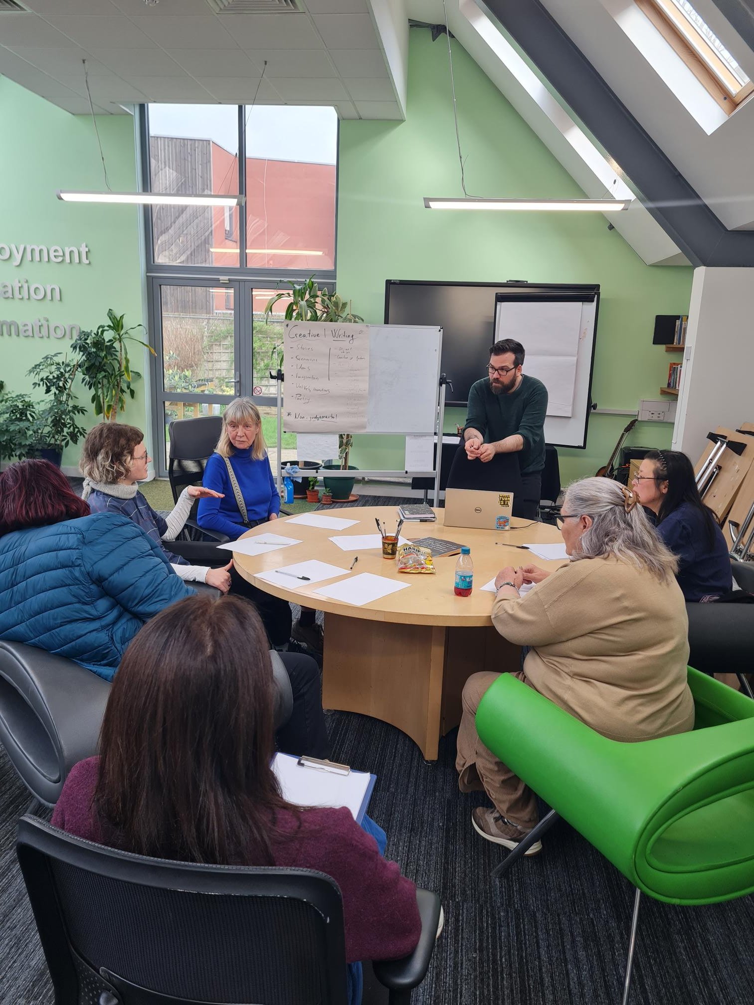 A group of diverse people attending a meeting in a modern conference room with green walls and large windows, seated around a round table with papers and drinks, and a presenter standing near a whiteboard.
