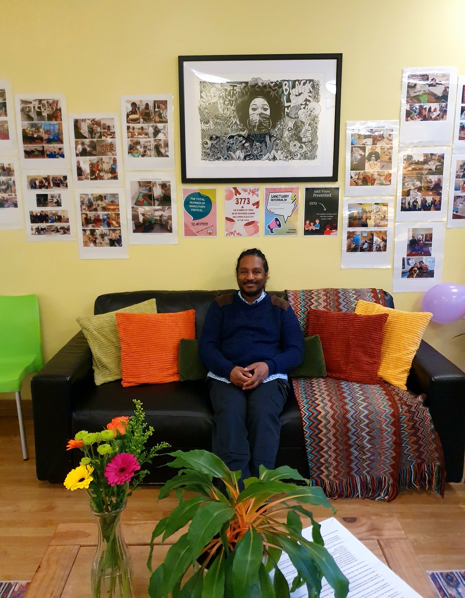 A man sitting on a black couch in a room decorated with colorful pillows, a photograph on the wall, and banners with various messages, with a vase of vibrant flowers in the foreground.