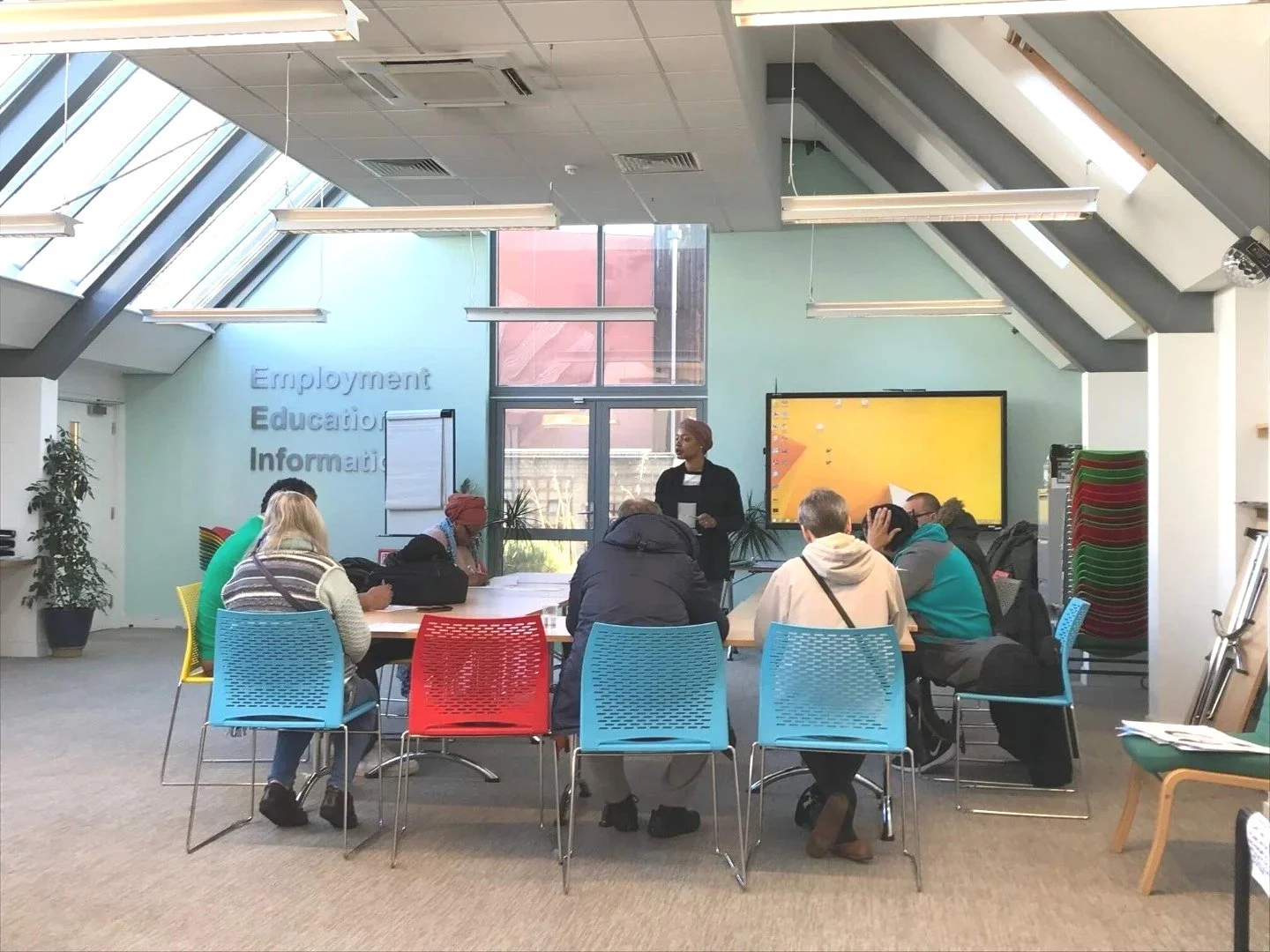 A diverse group of people seated around a circular table, attending a presentation in a modern, brightly lit room with large windows, colorful chairs, and a presenter standing in front of a large screen displaying a yellow background.