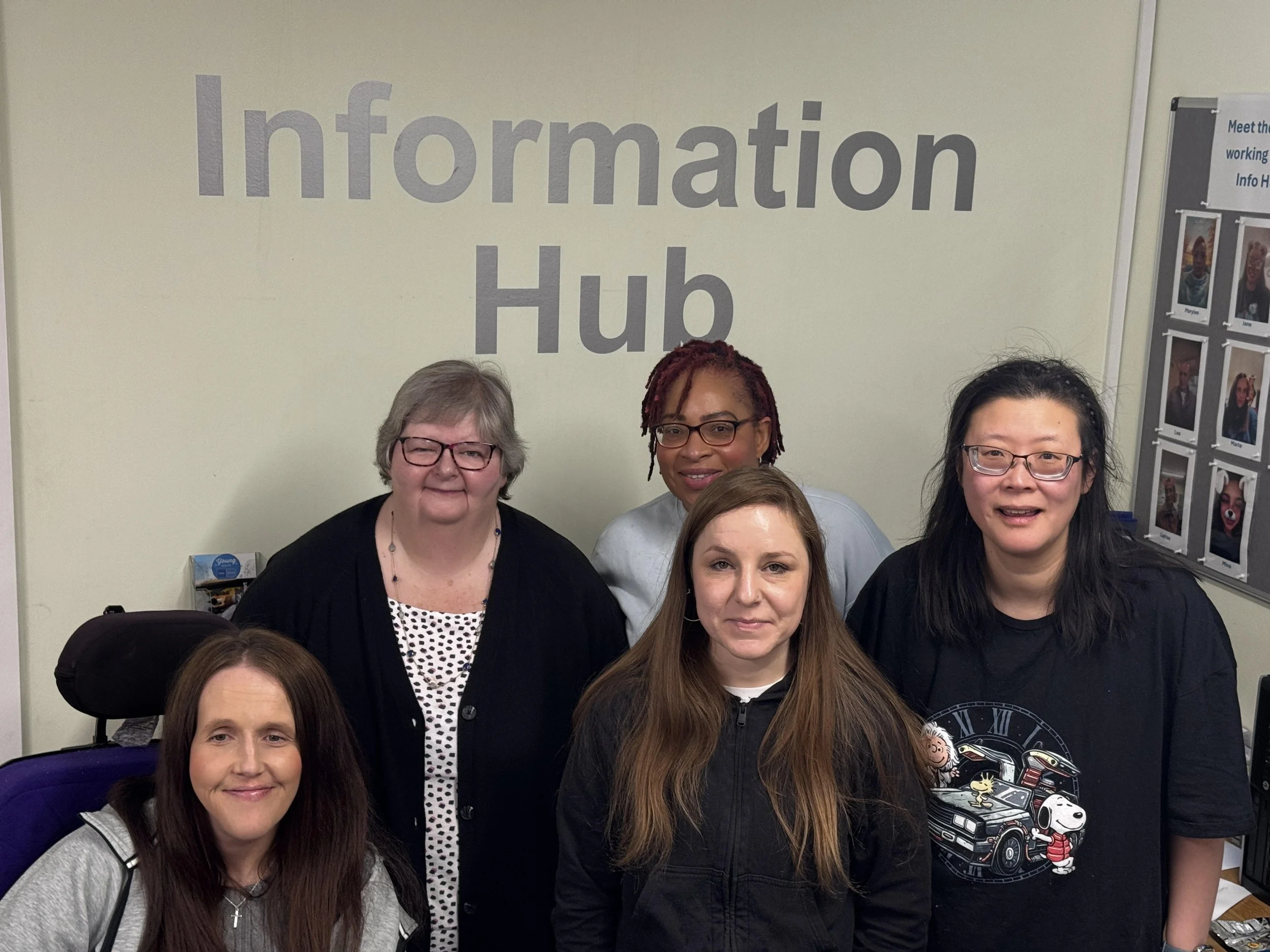 A group of women posing for a photo in front of an 'Information Hub' sign. The women are smiling and appear to be at a community or social event.