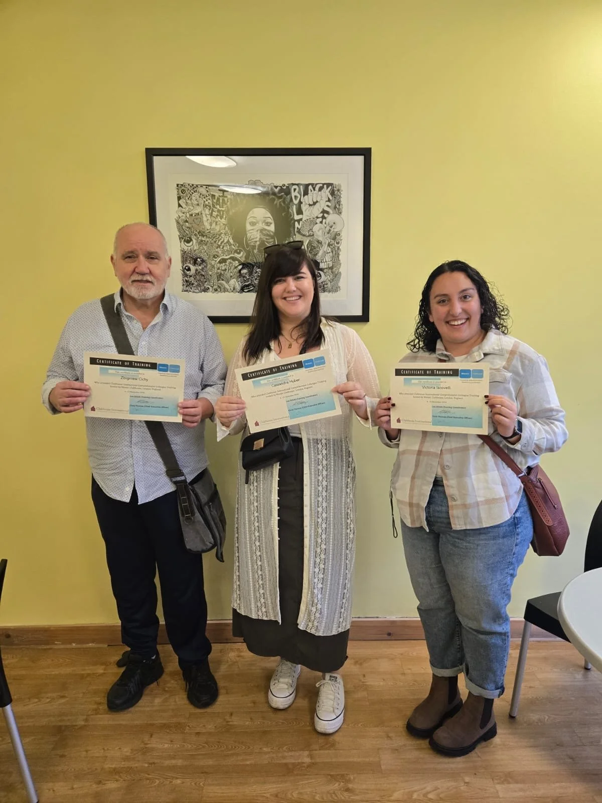 Three people standing indoors, each holding a certificate of training, smiling at the camera, with a framed black-and-white artwork on a yellow wall behind them.