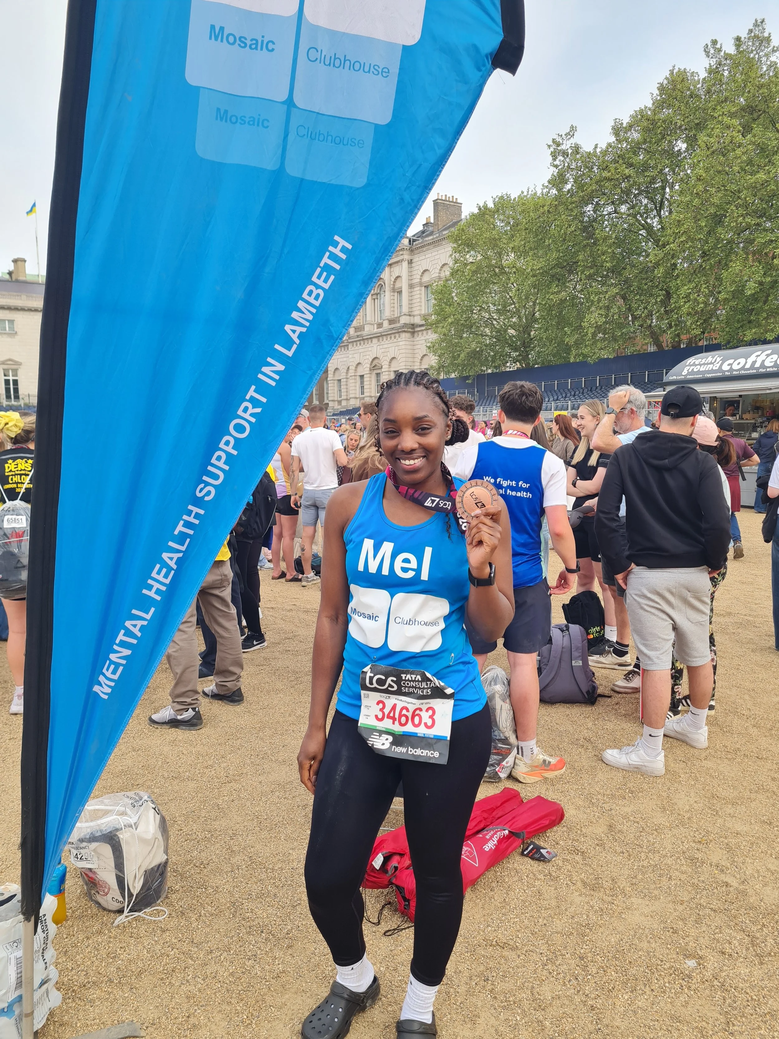 A young woman smiling and holding a medal at a marathon event, wearing a blue tank top with her name Mel, black leggings, and athletic shoes. There are other runners and spectators in the background. A blue flag with race support information and a race bib number 3463 are also visible.