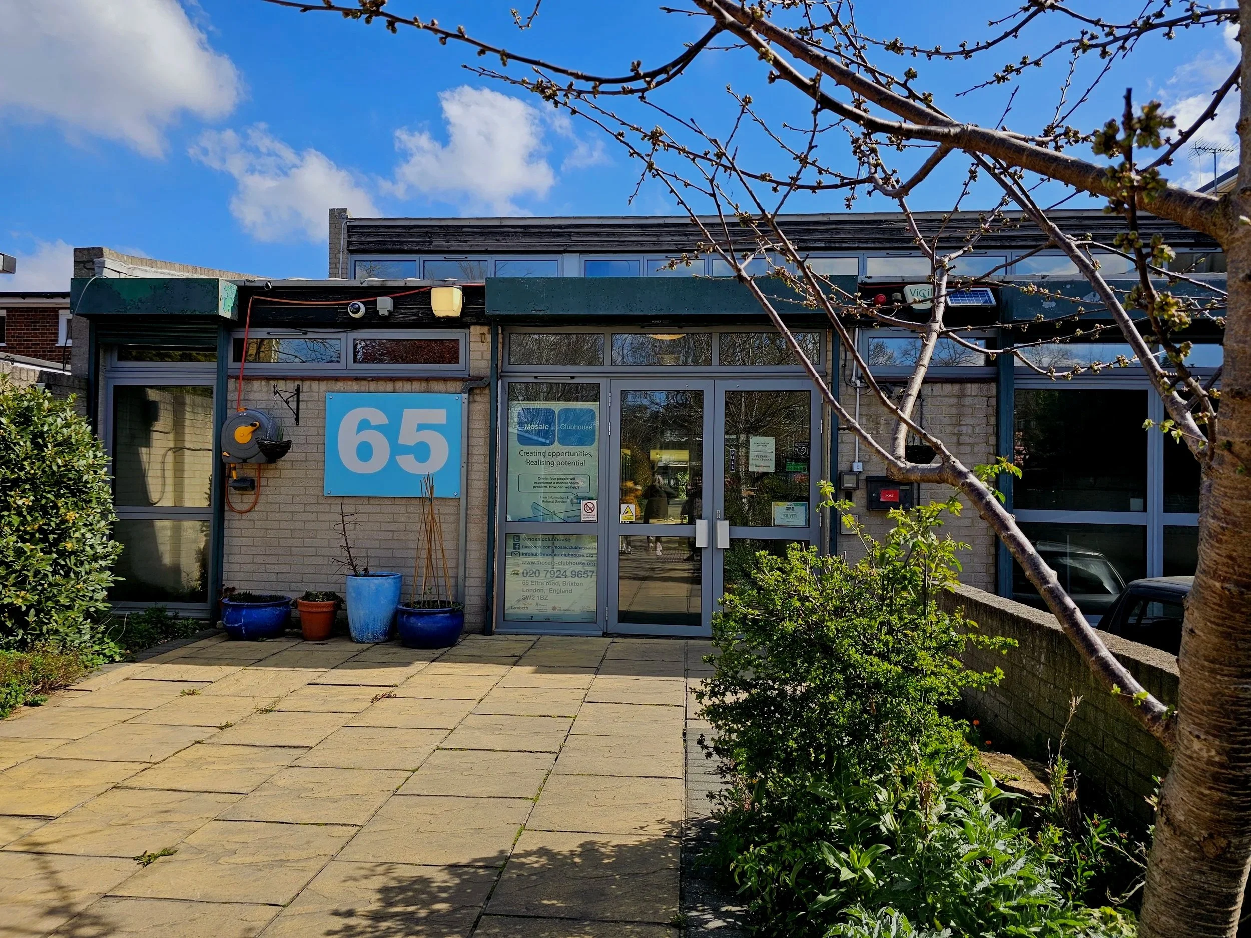 The front of a building with large glass doors and windows, a blue sign with the number 65, and a patio with potted plants. A tree with budding branches is visible in the foreground, and a blue sky with some clouds is above.
