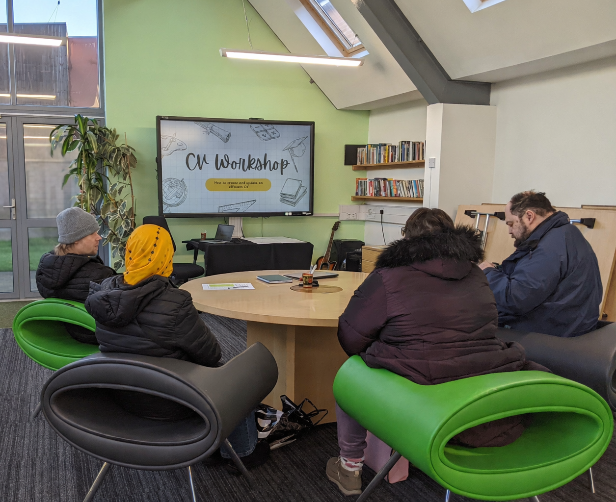 People attending a workshop in a modern classroom, sitting around a circular table, facing a large digital screen displaying 'CV Workshop' with illustrations and text about creating and updating a CV.