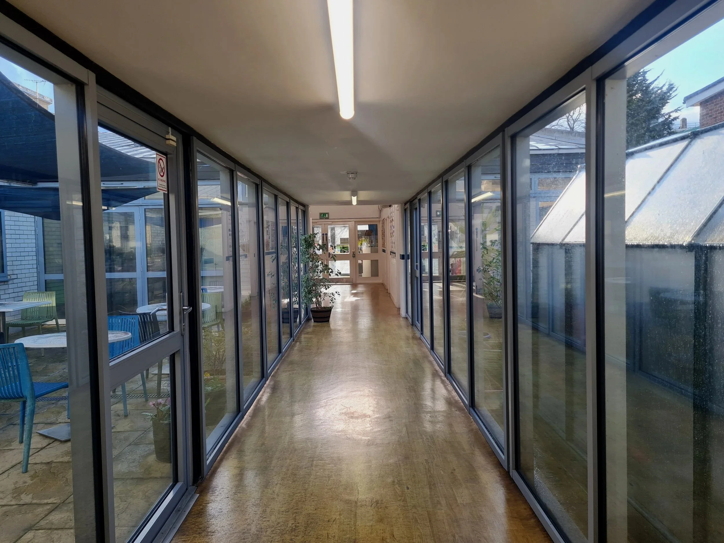 A covered walkway with glass walls and sliding doors leading to an outdoor patio with tables and chairs. Potted plants and ceiling lights are visible inside. Outside, there are trees and a brick building.