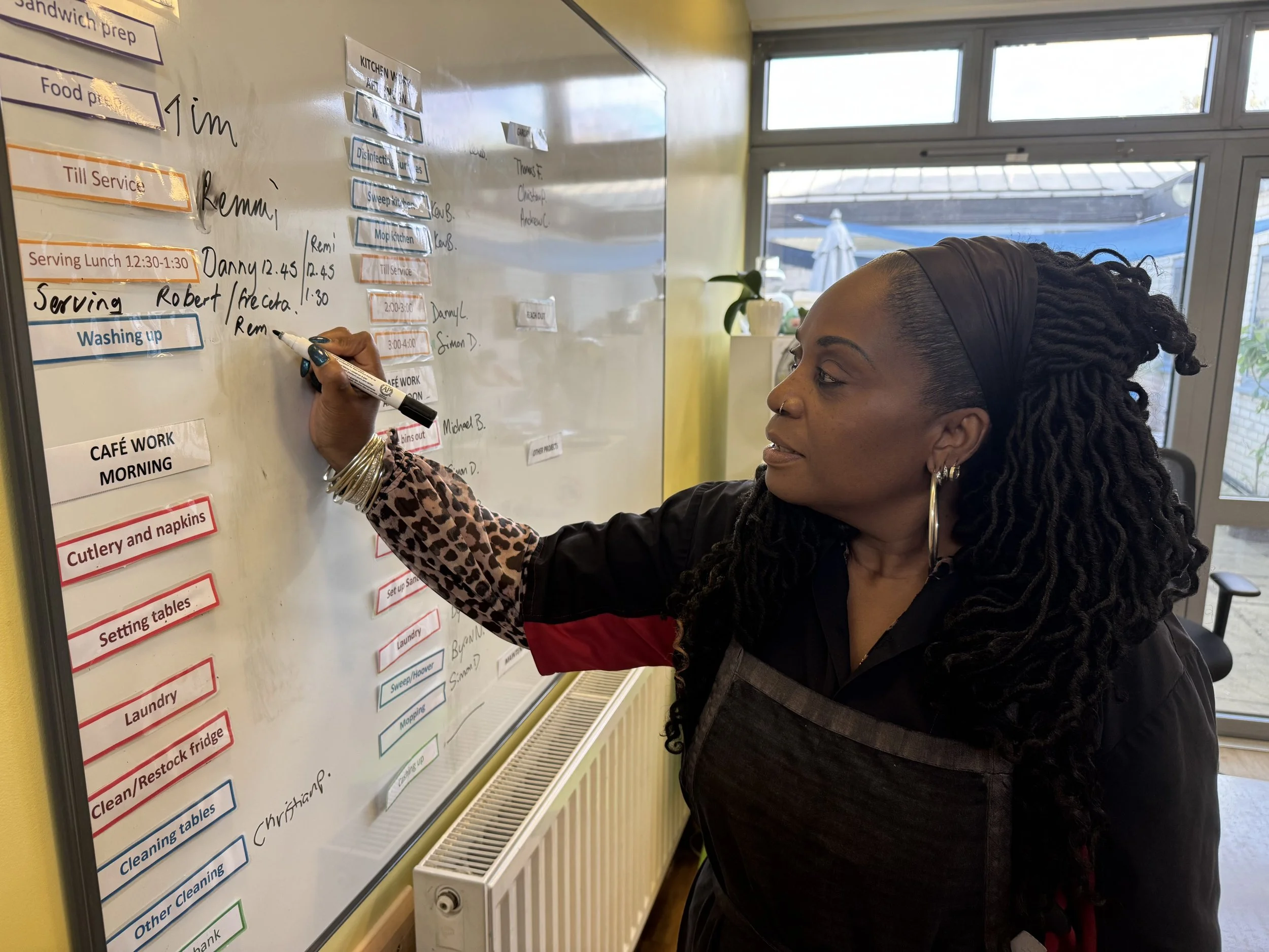 A woman with long dreadlocks wearing a black and leopard print top writes on a whiteboard with a black marker. The whiteboard contains a list of tasks related to kitchen and cleaning activities, with some items highlighted in orange and blue. The room has large windows showing an outdoor scene with a deck and umbrella, and a yellow wall with a radiator below the whiteboard.