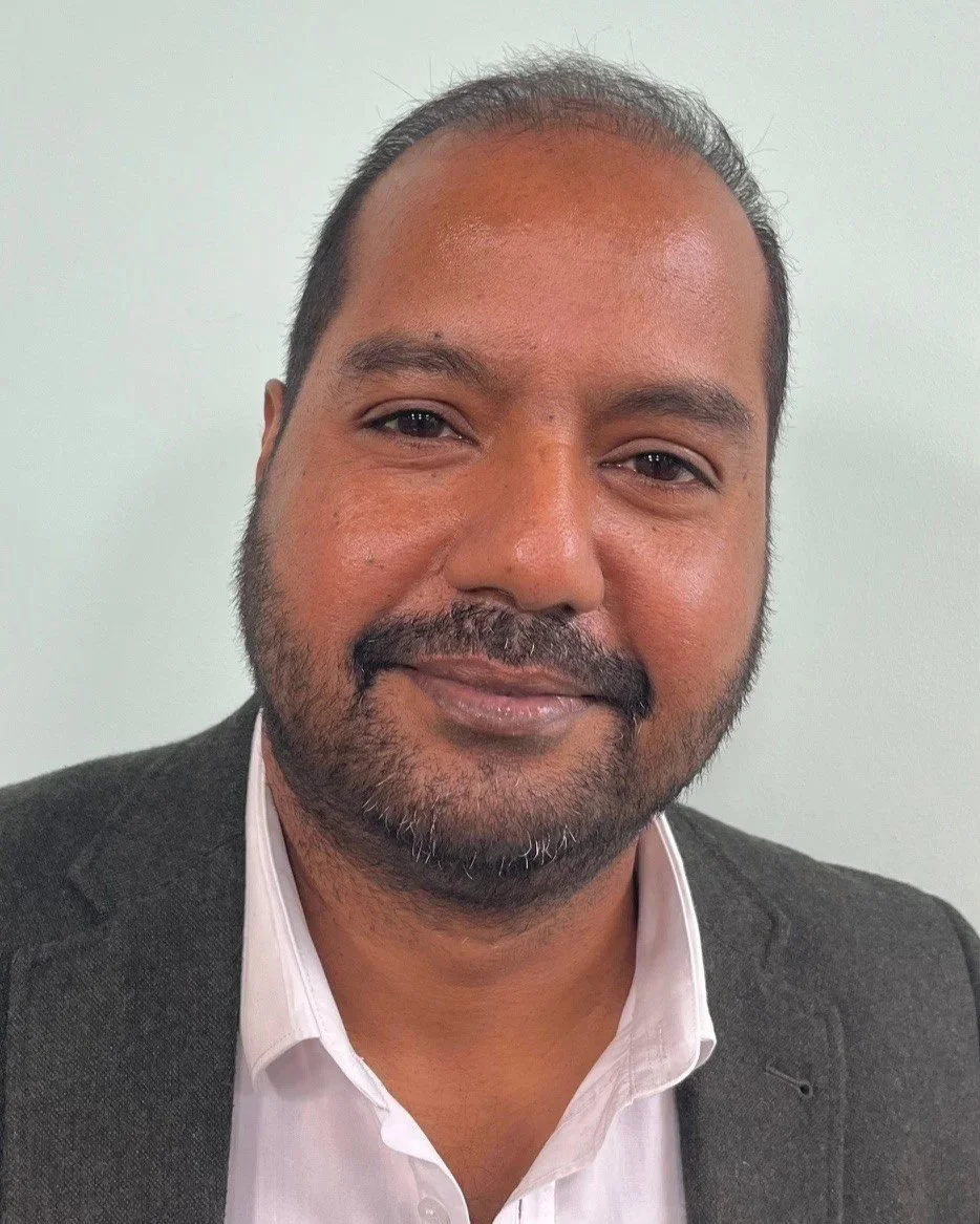 Headshot of a man with short hair and a beard, wearing a white shirt and dark blazer, smiling against a plain background.
