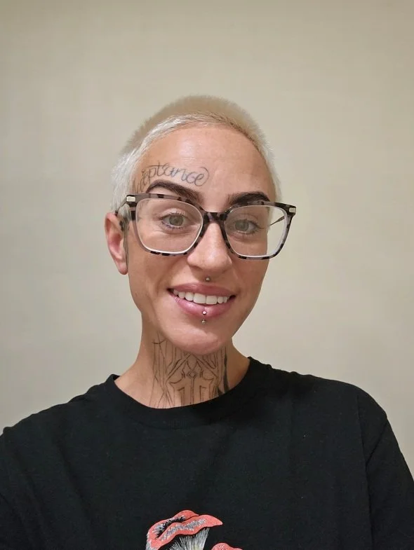 Person with short platinum blonde hair, glasses, tattoos on face and neck, smiling, wearing a black shirt with a red lips graphic.