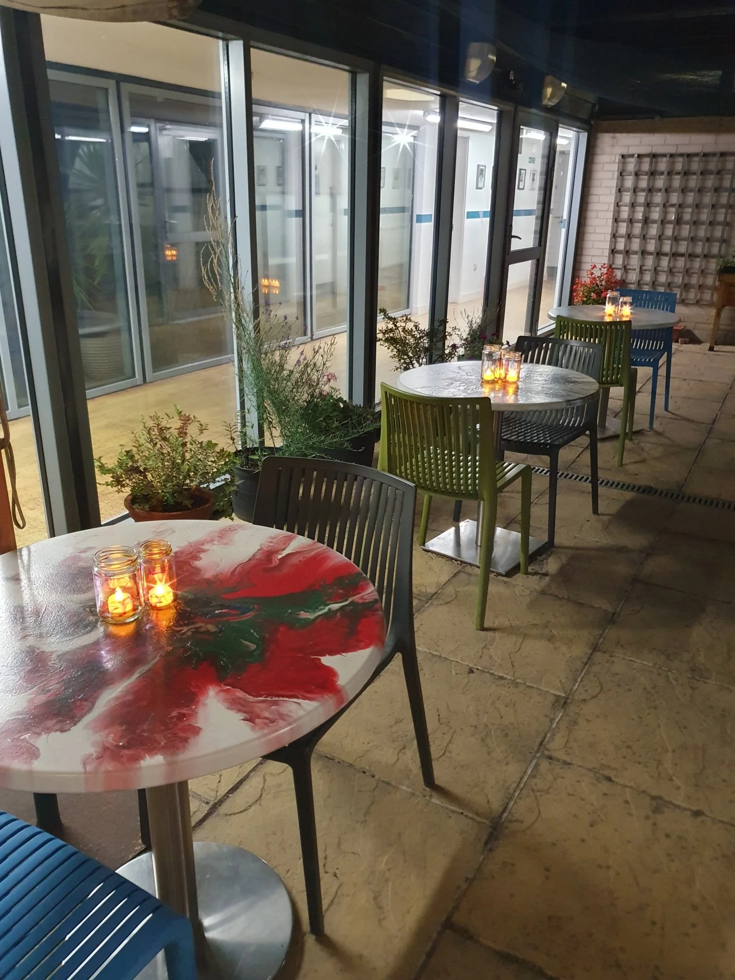 Empty outdoor patio with colorful plastic chairs and tables, decorated with candles, plants, and a brick wall, overlooking a building's interior through glass walls.