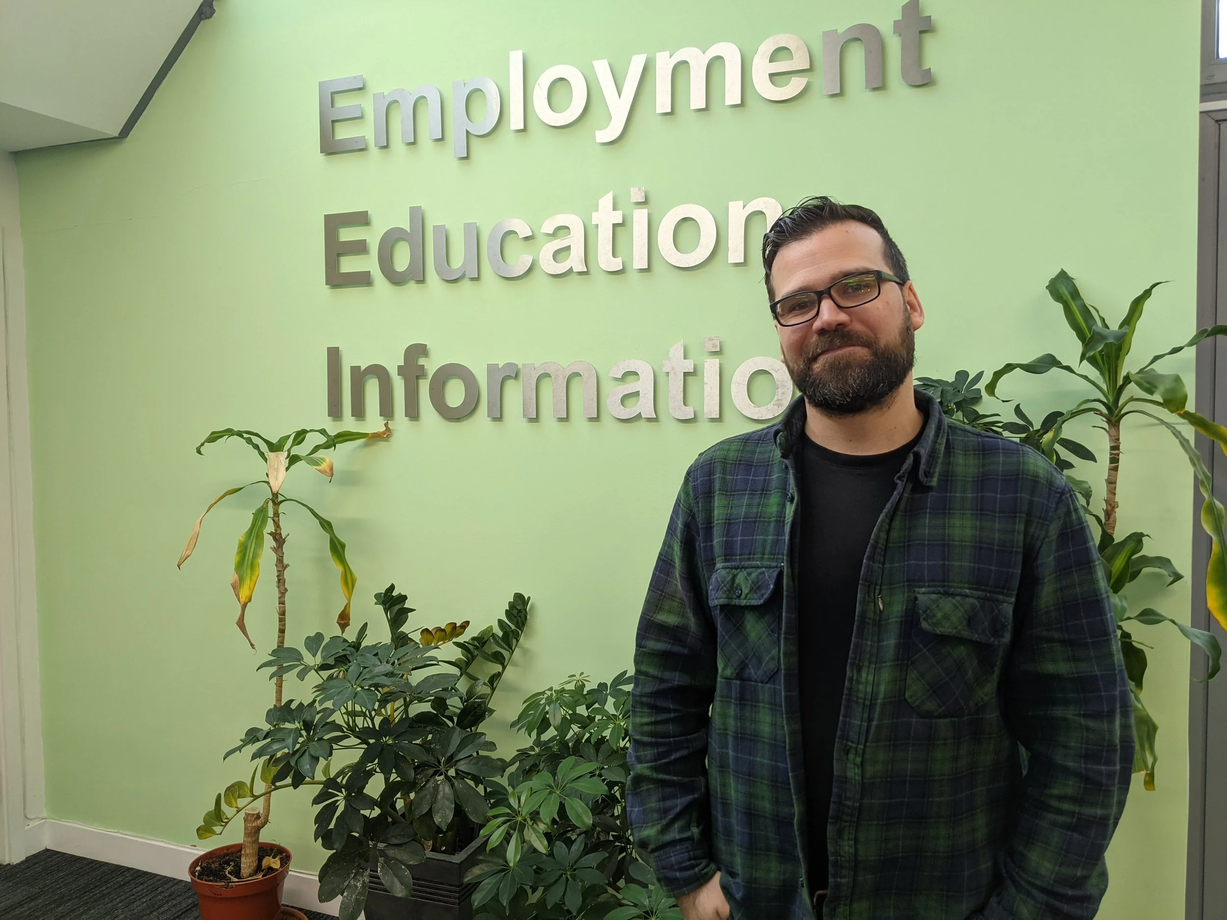 A man with glasses and a beard standing in front of a green wall with the words 'Employment', 'Education', and 'Information' in large letters. There are potted plants on either side of him.
