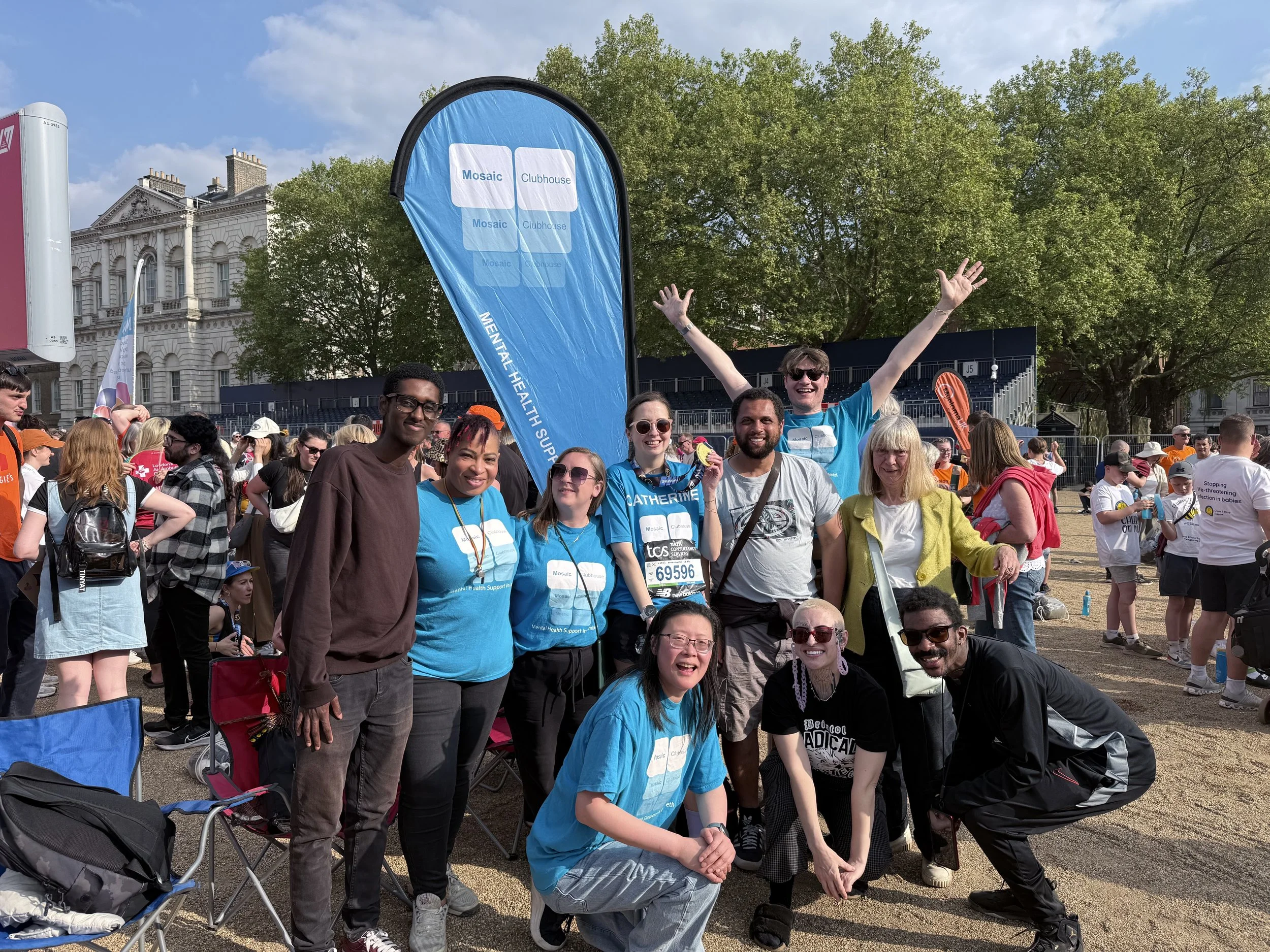 Group of diverse people at a community event outdoors with a blue flag that reads 'Mosaic Clubhouse' and 'Mental Health Support.' Some are smiling and posing for a photo, with tents, trees, and a historic building in the background.