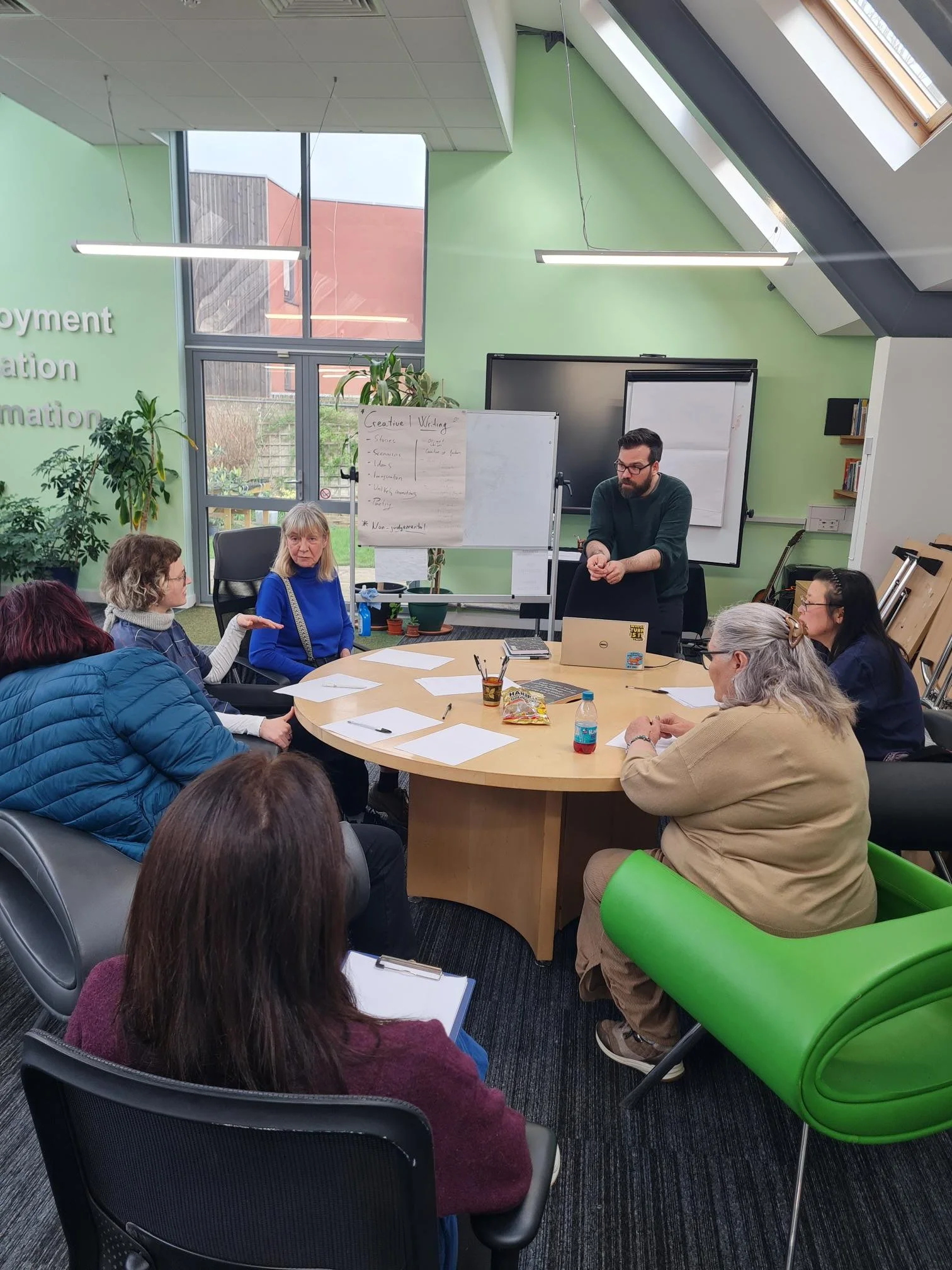 A group of women and a man participating in a meeting or workshop in a modern conference room with green walls, large windows, and a round table, with notebooks, papers, and snacks on the table.