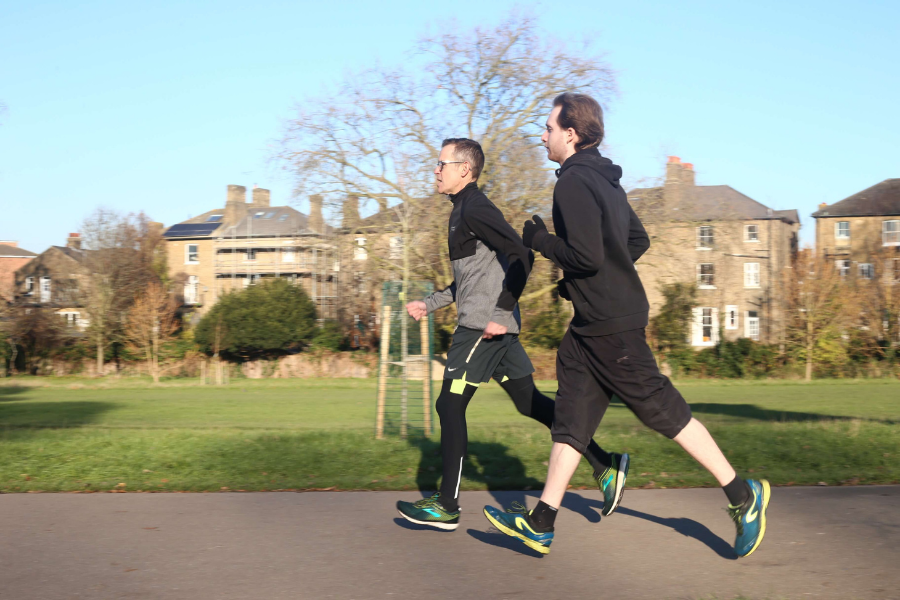 Two men running outdoors on a paved path in a park, with grass, trees, and residential buildings in the background.
