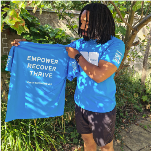 Person with dreadlocks wearing a blue T-shirt holding up another blue T-shirt with the words "EMPOWER RECOVER THRIVE" printed on it, outdoors with greenery in the background.