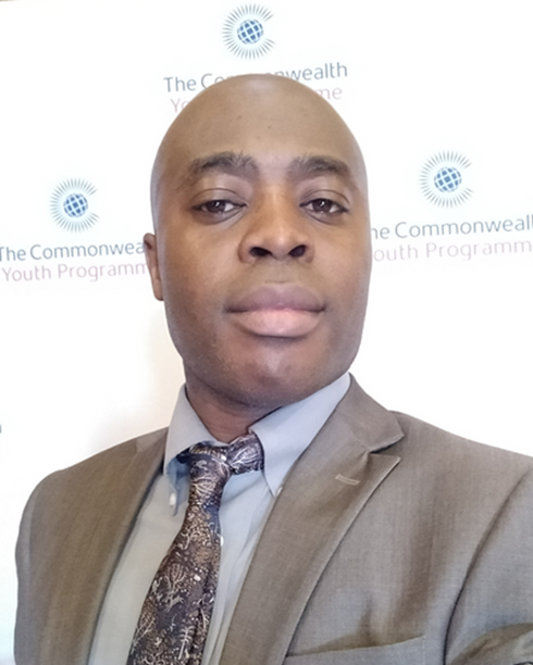 A man wearing a gray suit and patterned tie taking a selfie in front of a backdrop with the Commonwealth Youth Programme logo.