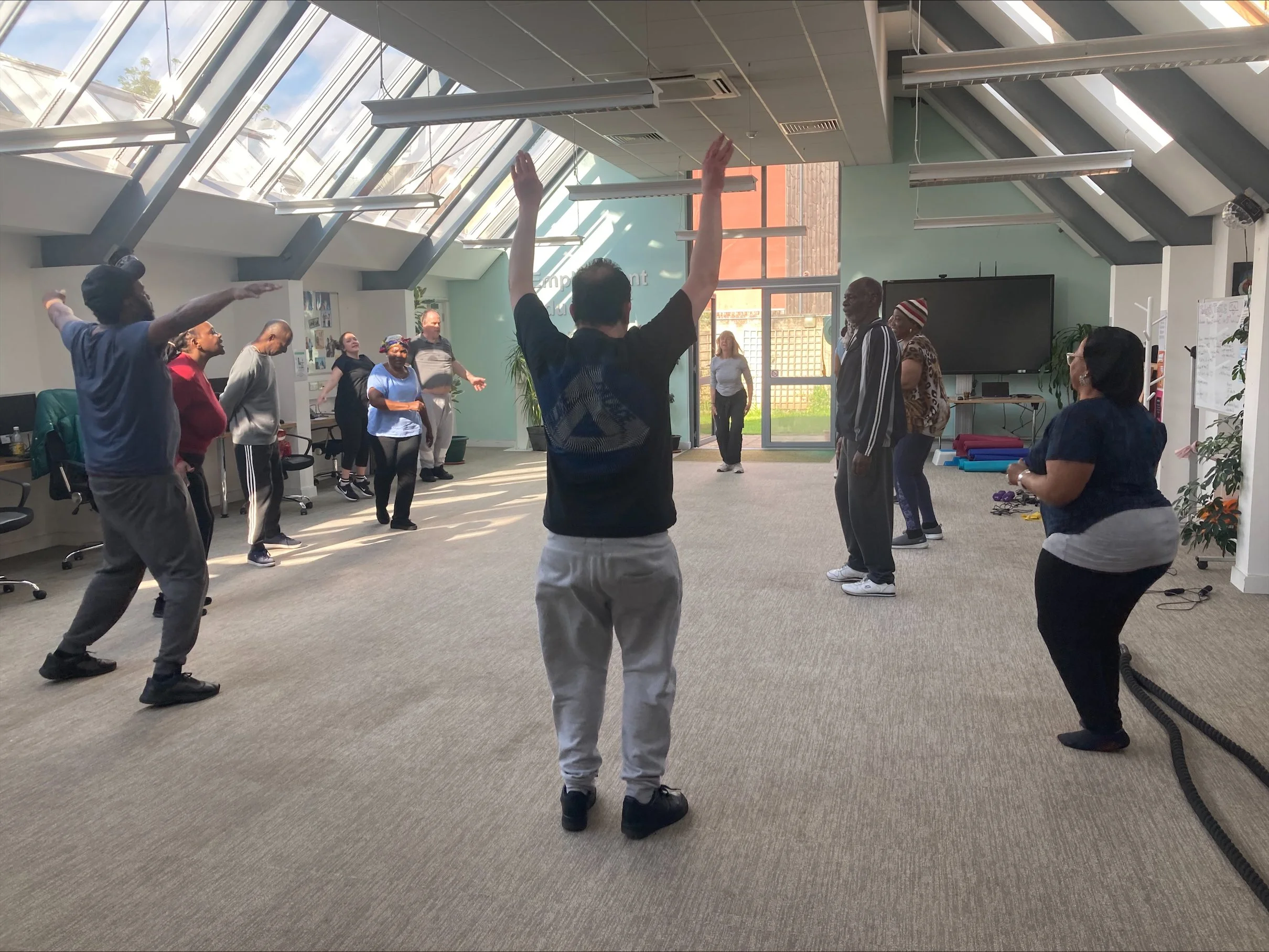 A diverse group of people participating in a fitness class in a bright room with sunlit skylights, some with arms raised, following an instructor at the front.