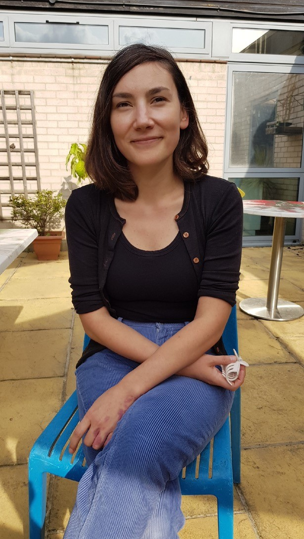A woman with shoulder-length brown hair, wearing a black top and a black cardigan, sitting outdoors on a blue chair, smiling, with potted plants and a building with brick walls and glass windows in the background.
