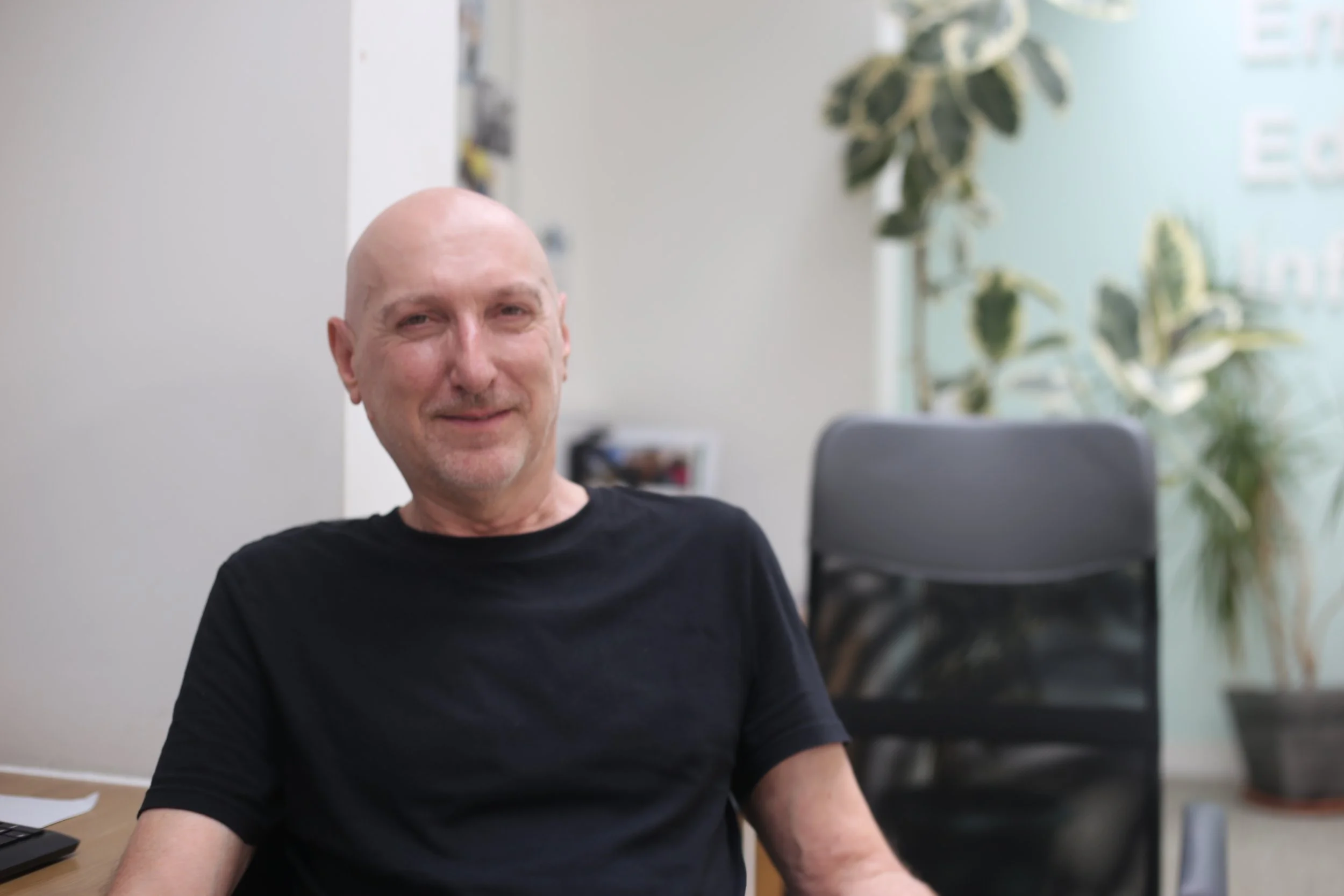 A smiling bald man in a black T-shirt sitting in an office with potted plants and a black chair in the background.
