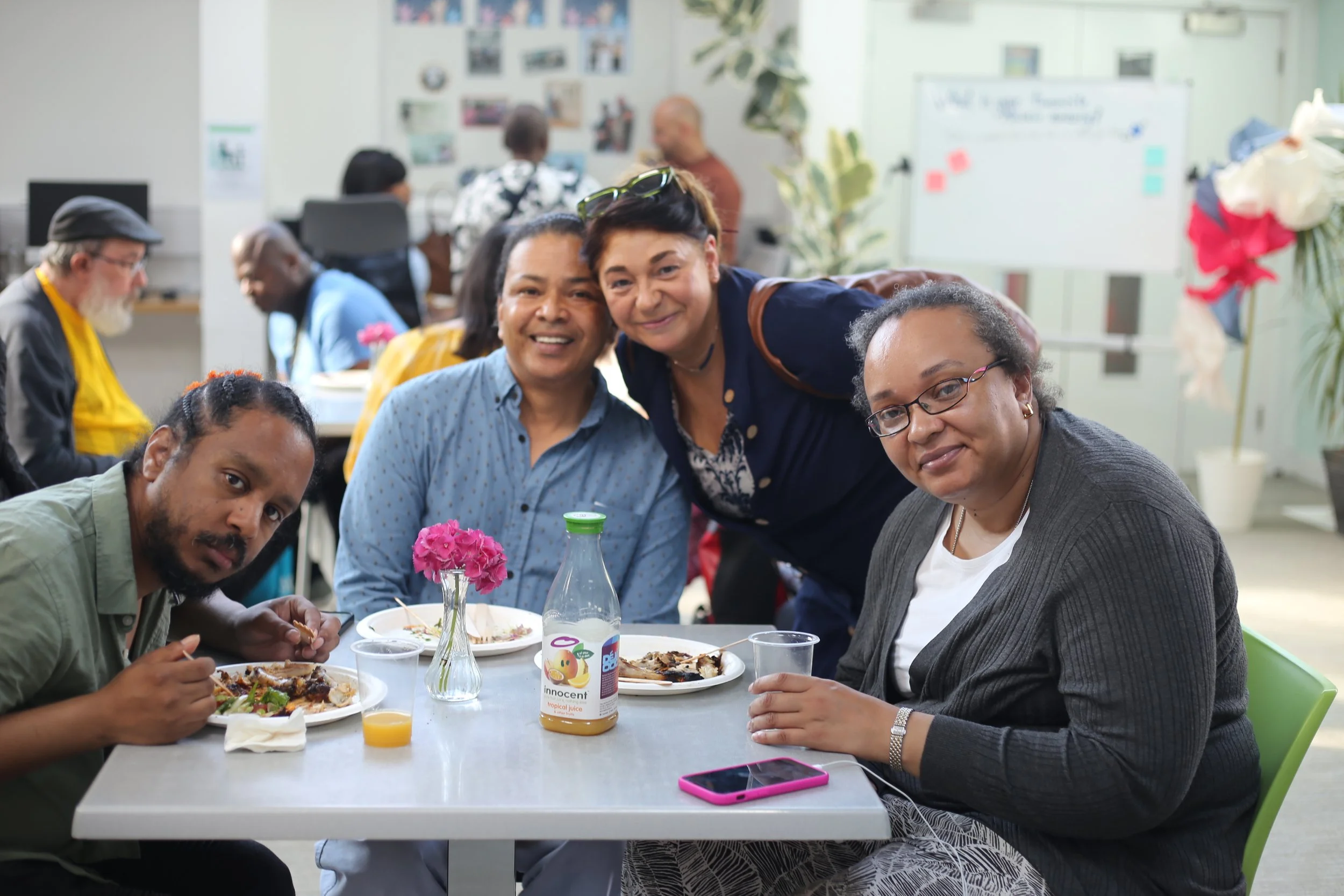 A diverse group of people sitting at a table enjoying a meal in a bright, decorated indoor space. Some are smiling at the camera, and others are engaged in eating or conversation. There are flowers, plates of food, and drinks on the table, with a background featuring people working and a whiteboard on the wall.