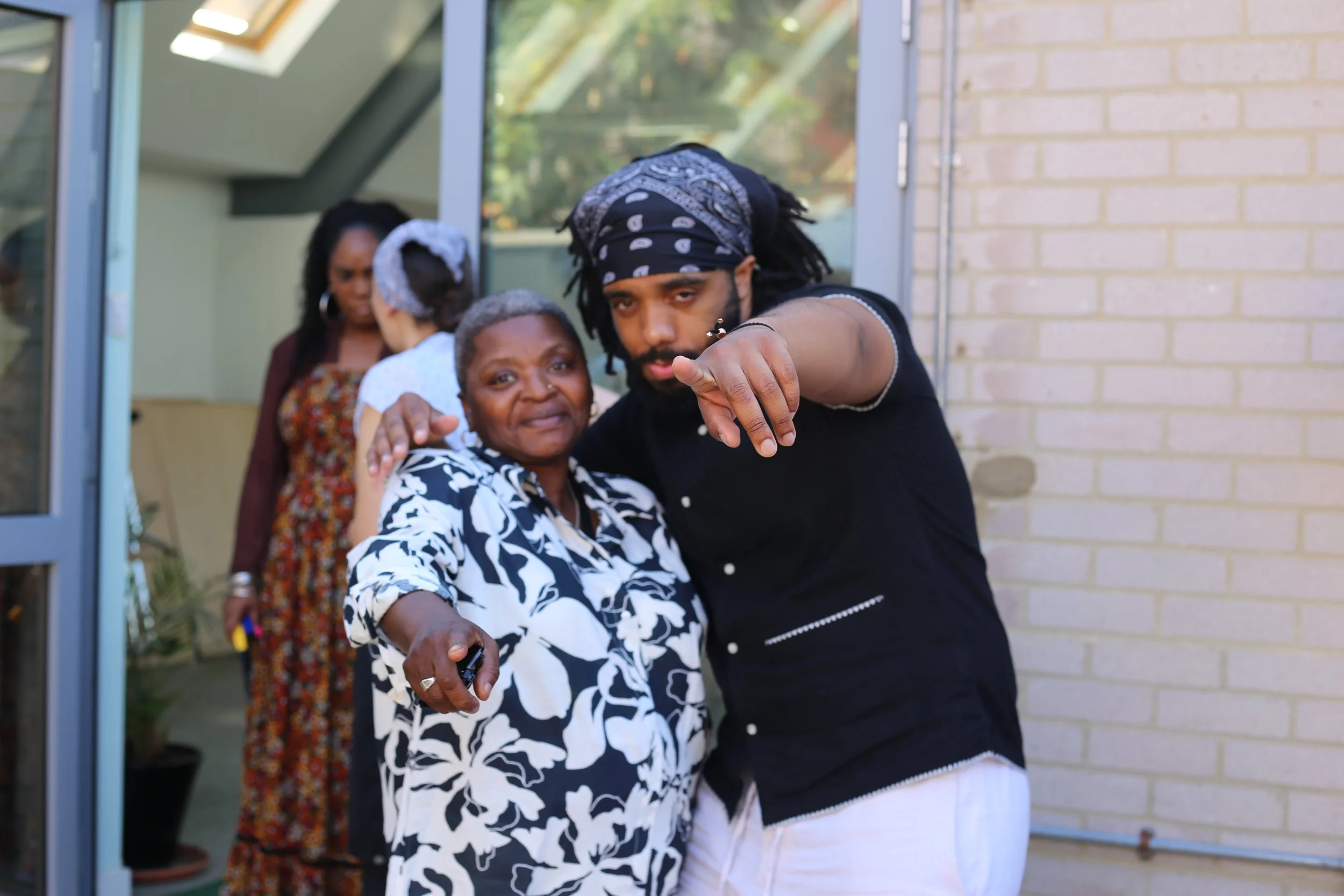 A young man with a bandana and earrings points towards the camera while embracing an older woman in a black and white floral shirt. Behind them, three women are conversing near an open door, one of whom is wearing a floral dress and headscarf. The setting appears to be outside a house.