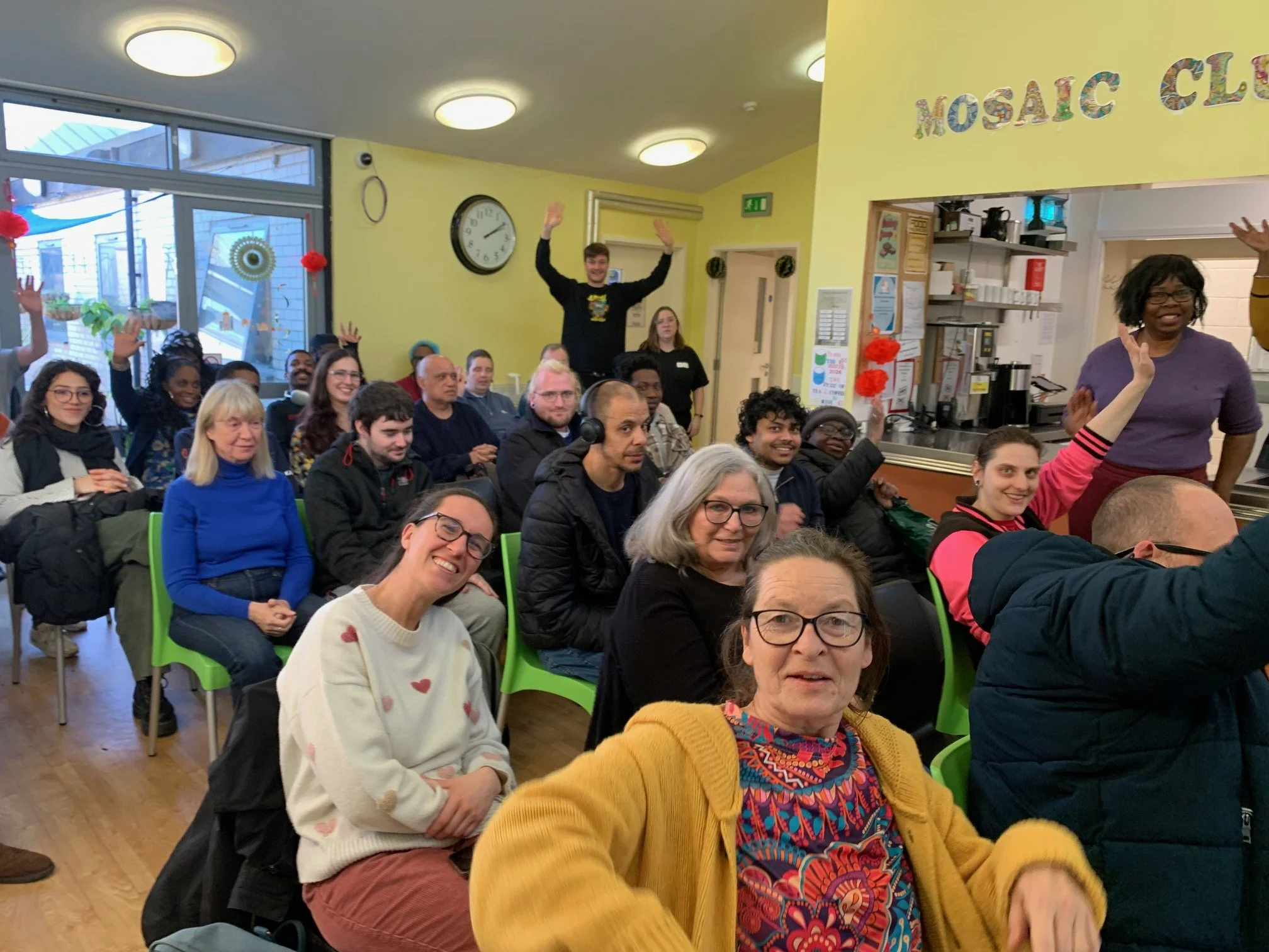People sitting and standing in a room during a group event or meeting, some raising their hands and smiling, with a yellow wall and a sign that reads 'MOSAIC CLUB' in colorful letters.