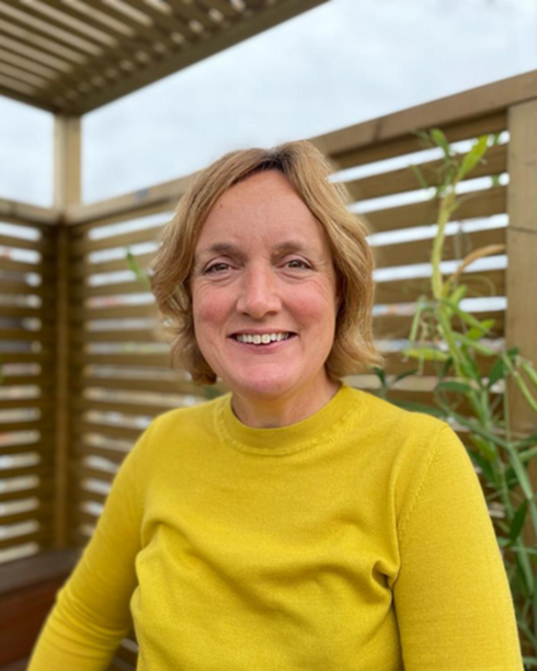 A woman with short blonde hair smiling, wearing a yellow long-sleeve top, standing on a wooden balcony with plants and a cloudy sky in the background.