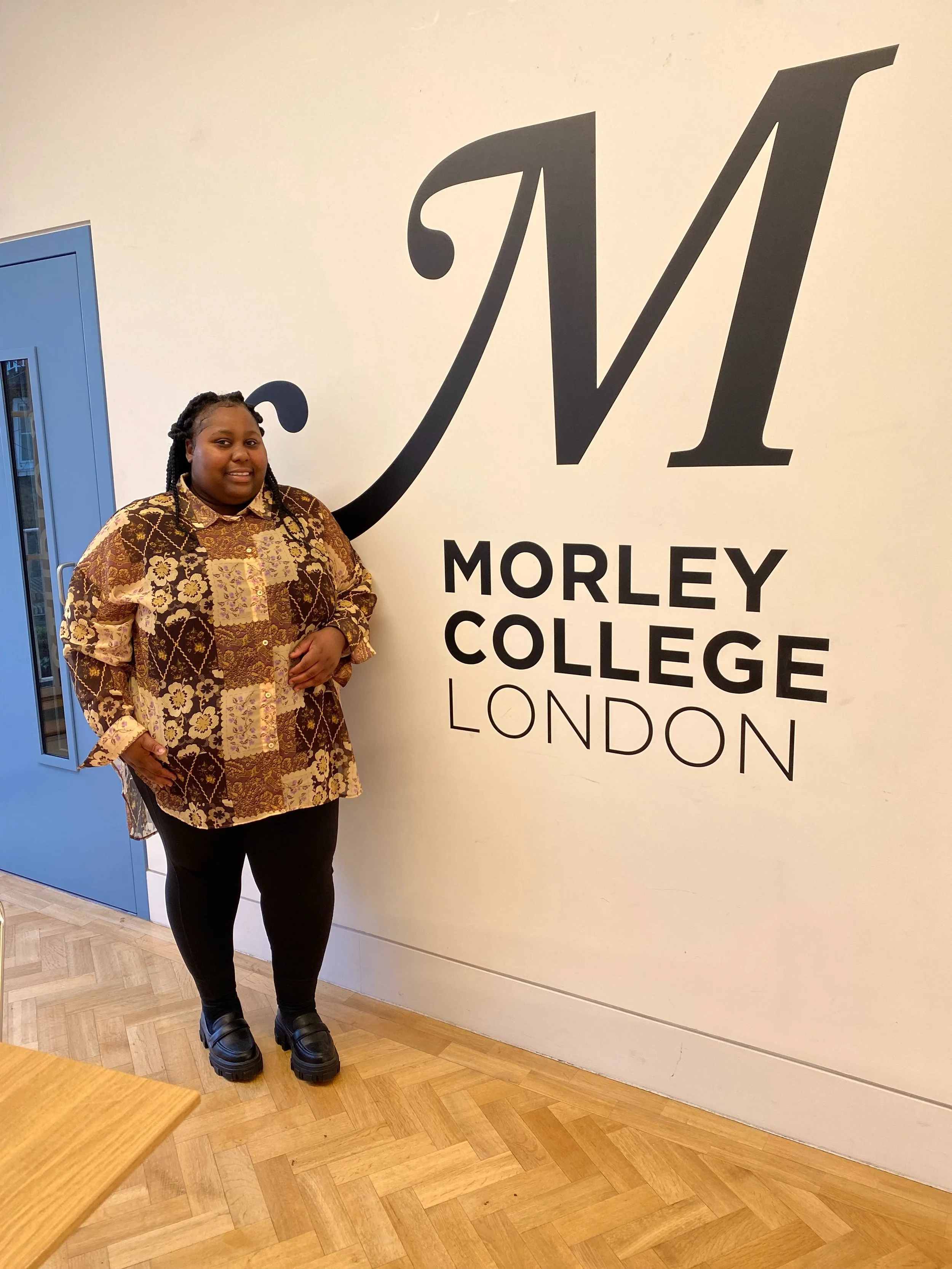 A woman standing indoors next to a large wall sign reading "Morley College London" on a white wall. She is wearing a patterned brown and yellow shirt, black pants, and chunky black shoes.