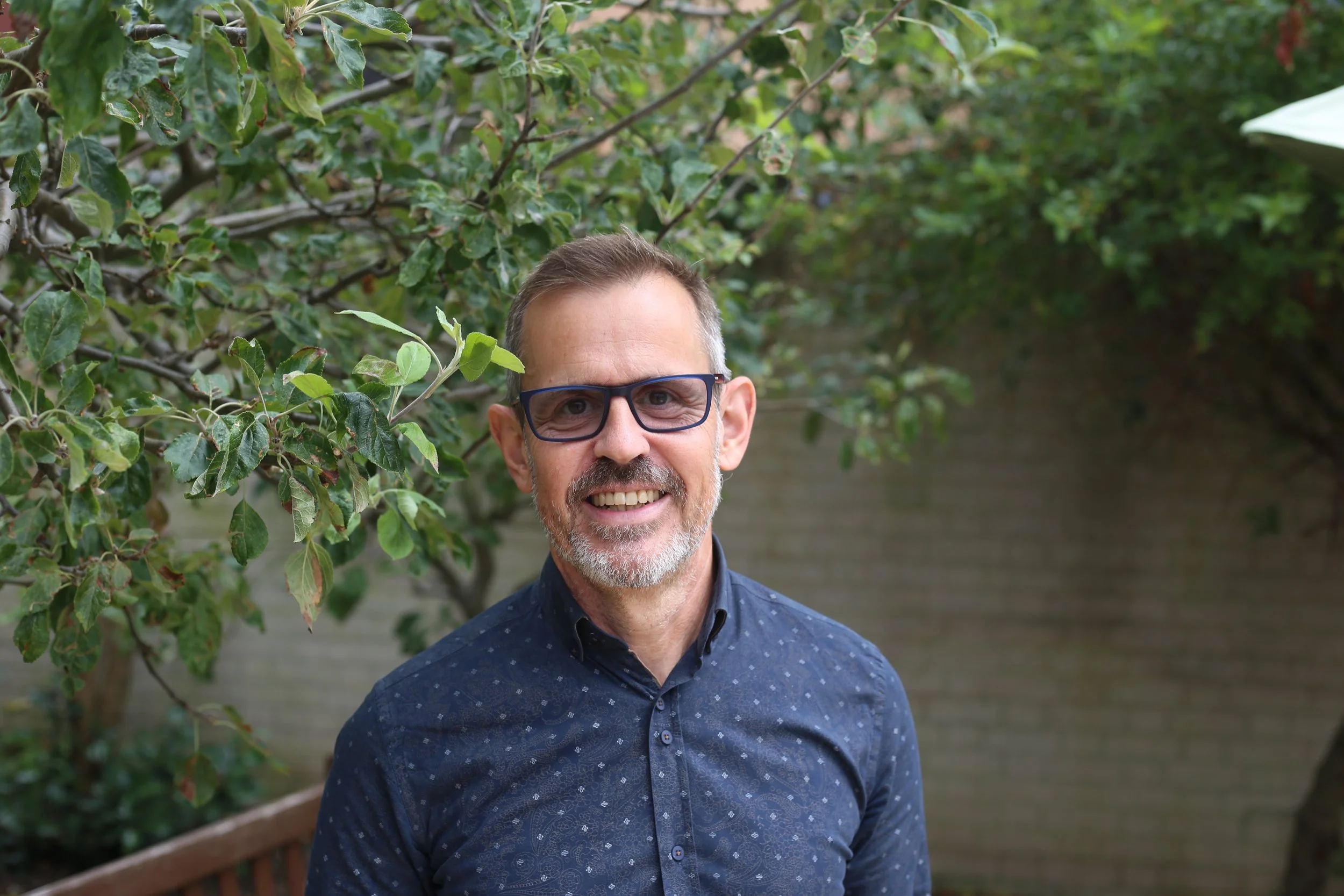 A middle-aged man with glasses, a beard, and short hair smiling outdoors, standing in front of green foliage and a brick wall.