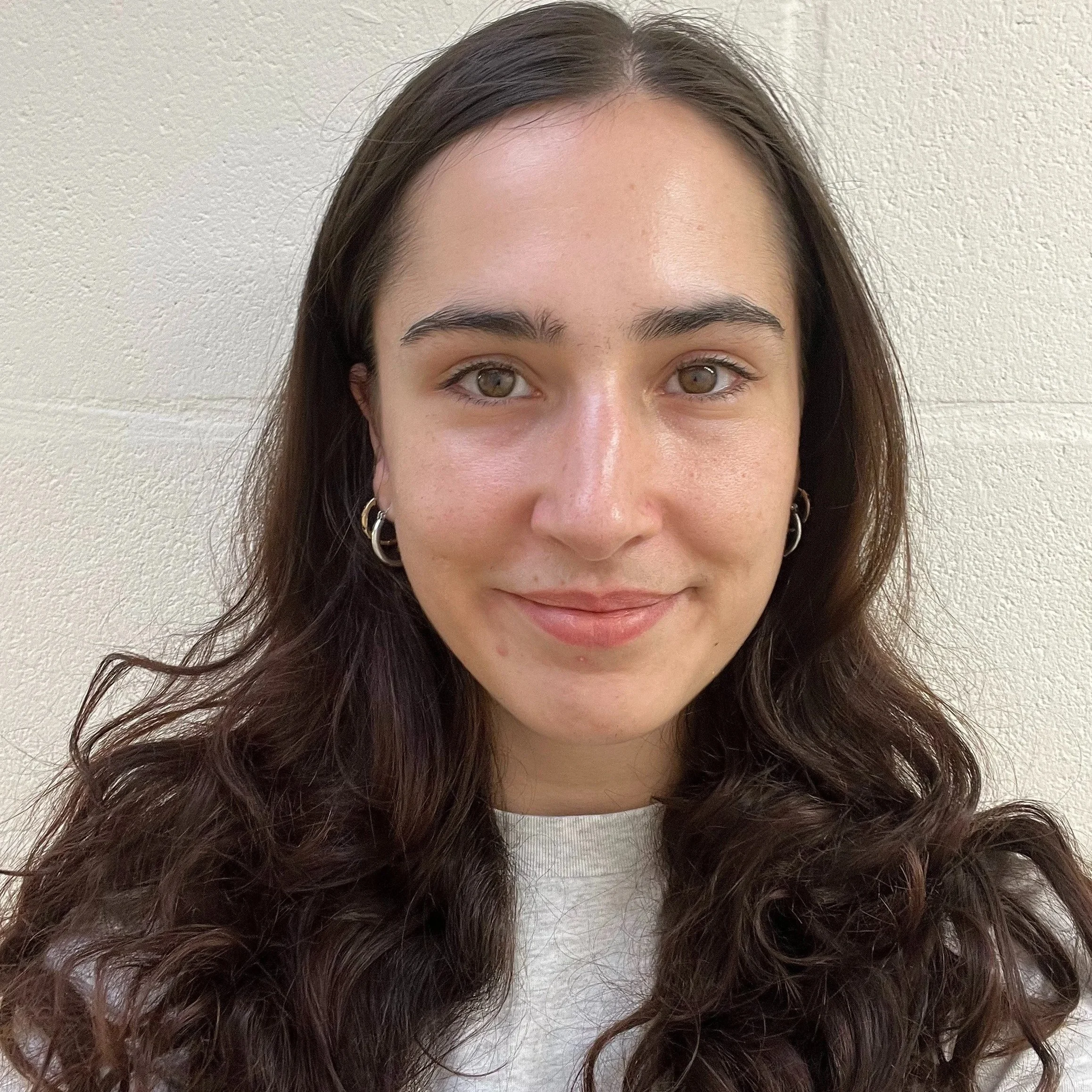 Close-up of a young woman with long wavy brown hair, hoop earrings, and wearing a light-colored top, standing against a plain beige wall.