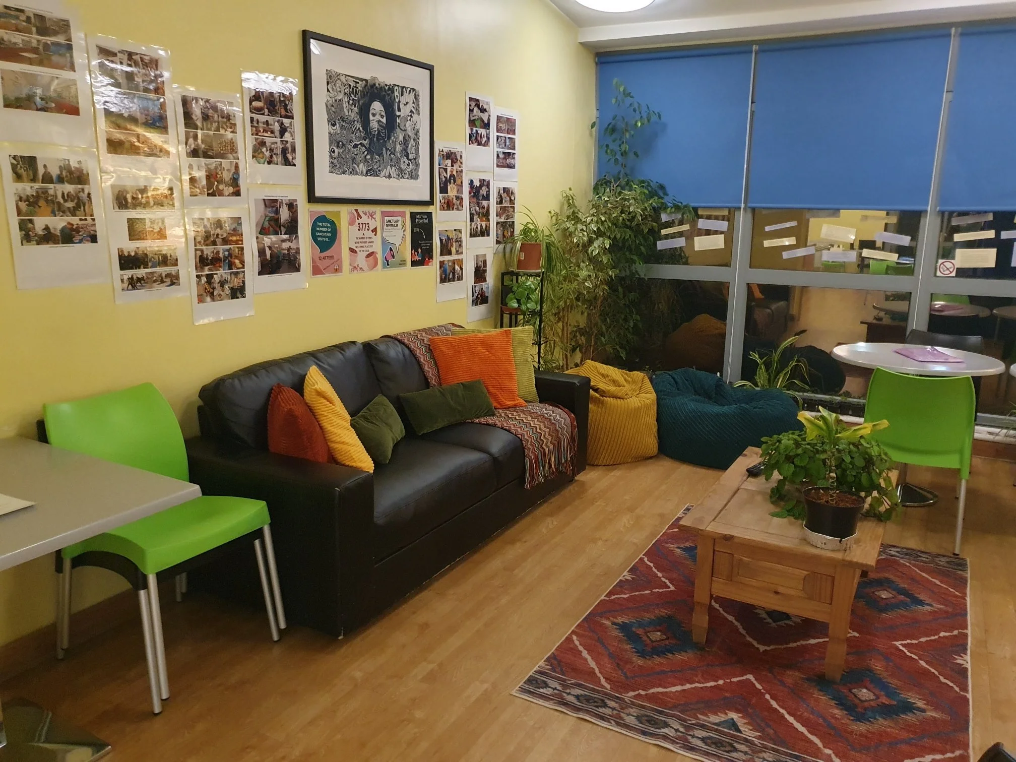 Cozy room with yellow walls, black sofa with colorful pillows, green chairs, and a wooden coffee table on a patterned rug. Wall decorated with photos and artwork. Plants near large window with blue blinds.