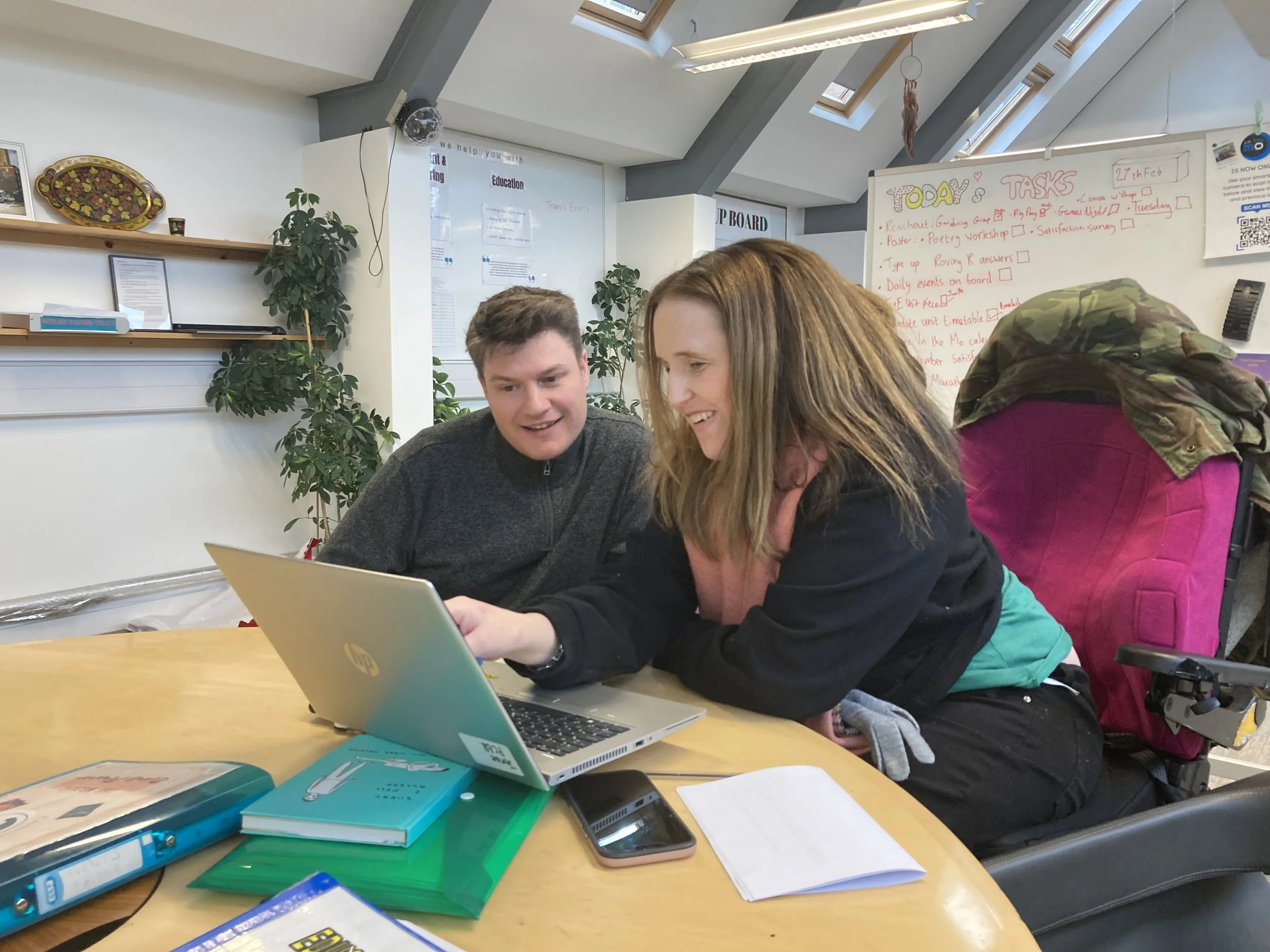 Two people, a young man and a young woman, are sitting closely together at a table working on a laptop. The woman is wearing a colorful jacket and the man is wearing a dark sweater. The table has notebooks, papers, a smartphone, and a blue binder. There is a whiteboard behind them with tasks written on it, and the room has large skylight windows and several green plants.