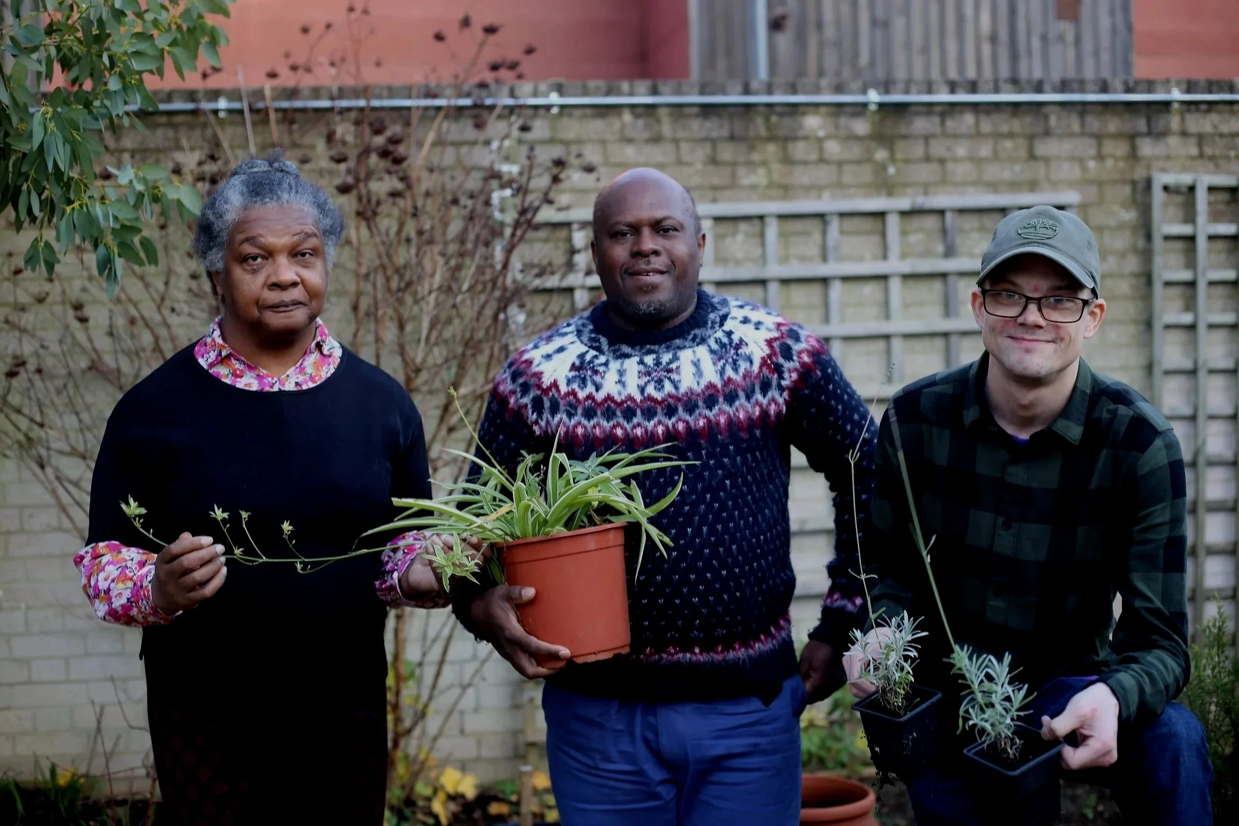Three people standing outside in a garden or backyard, posing for a photo. The person on the left is an elderly woman with gray hair pulled back, wearing a black sweater over a floral shirt. The person in the middle is a bald man with a patterned sweater, holding a potted plant with green leaves. The person on the right is a young man with glasses and a cap, wearing a dark plaid shirt, holding a smaller potted plant with light purple or grayish leaves.