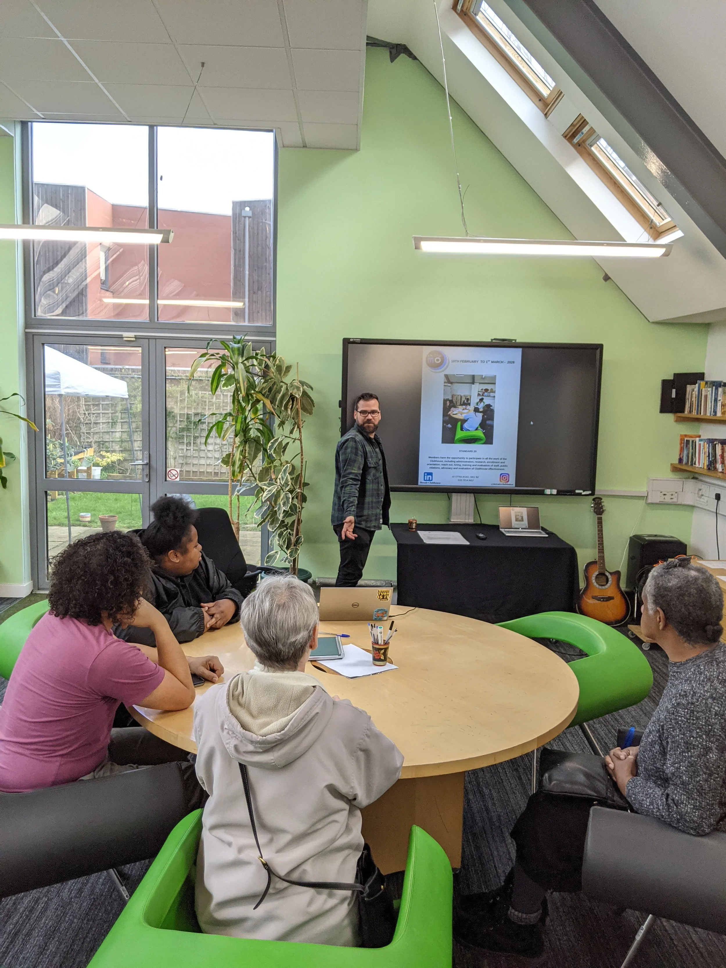 A diverse group of people sitting around a wooden table in a bright room, listening to a man standing next to a large screen displaying a presentation. The room has green walls, a large window, and skylights.