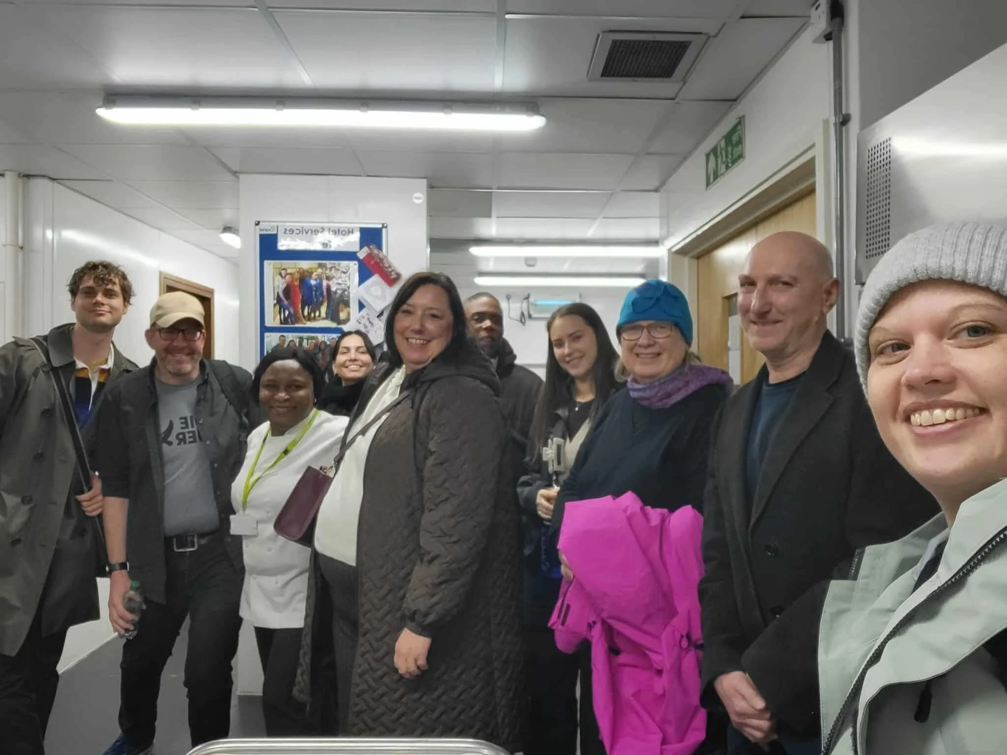 A group of diverse people smiling and posing for a photo indoors, possibly in a hospital or clinic hallway.