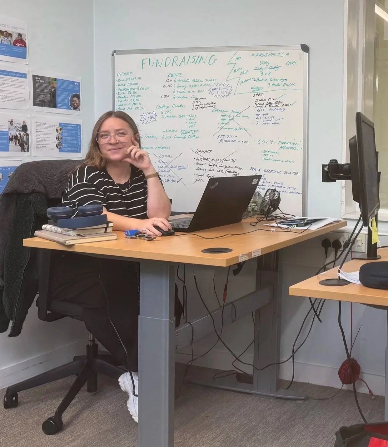A woman wearing glasses and a black and white striped shirt sitting at a desk with a laptop, notebooks, and a mouse, in front of a whiteboard with notes about fundraising and events, in an office.