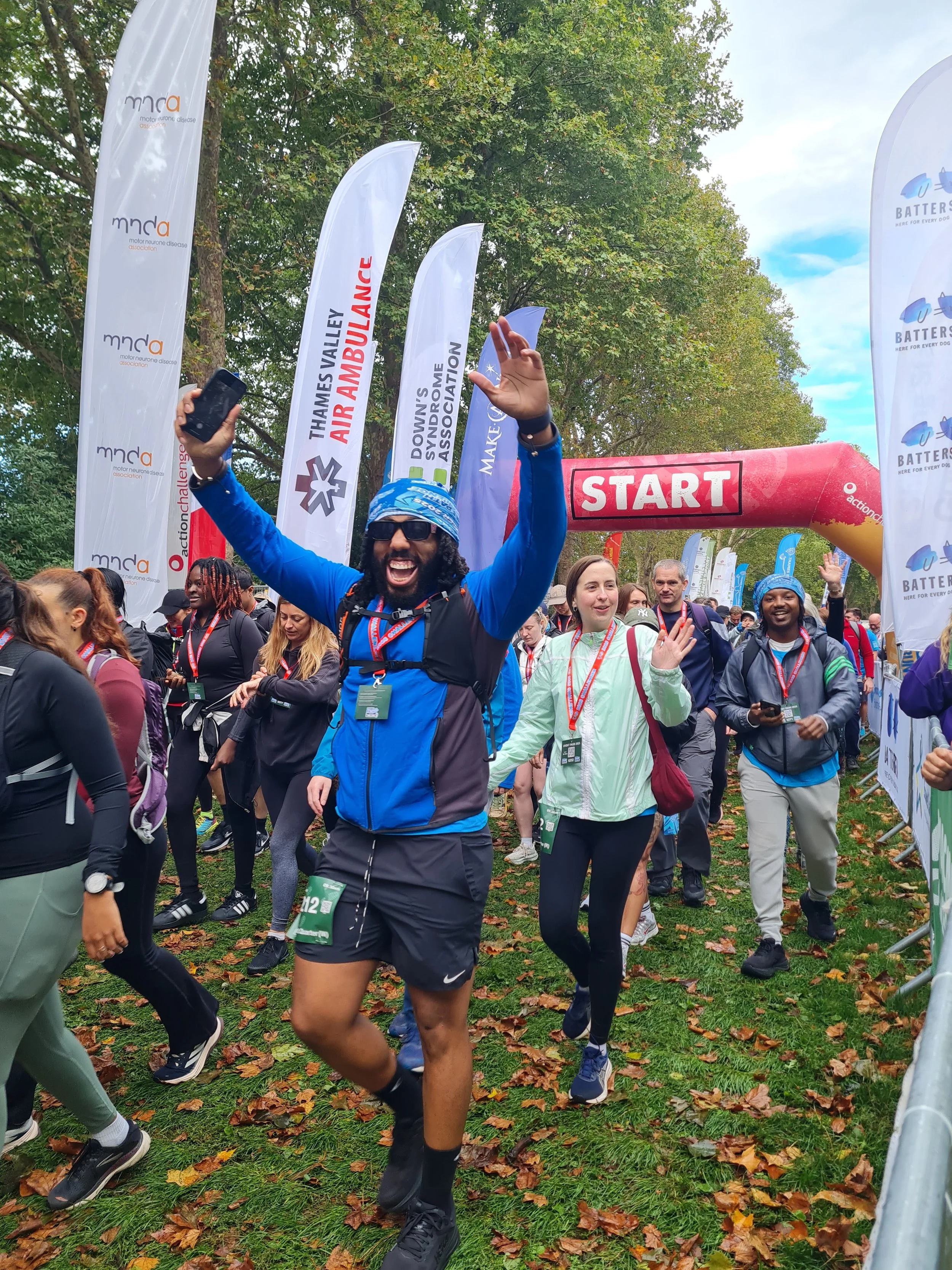 Group of runners at the start line of a race in a park, with trees and leaves on the ground, and a red start banner overhead, some people smiling and celebrating.