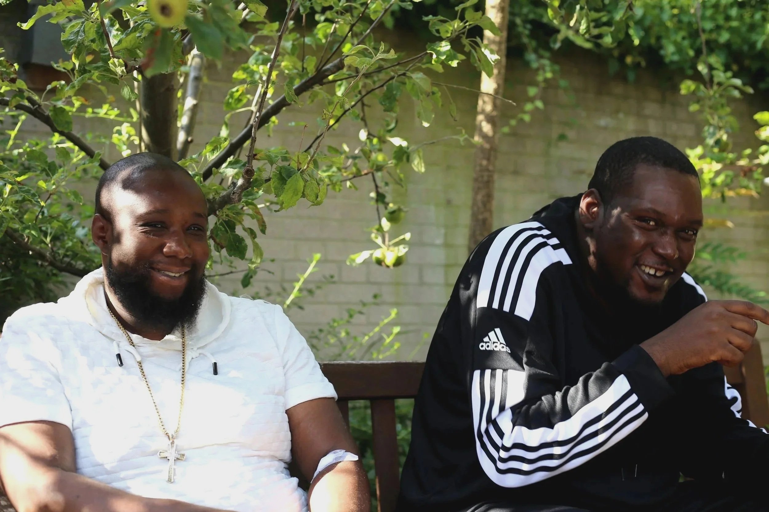 Two smiling men sitting on a wooden bench outdoors, one wearing a white shirt with a cross necklace, and the other wearing a black Adidas jacket, with green foliage and a brick wall in the background.