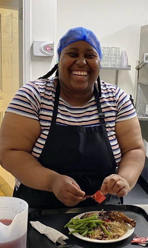 A woman wearing a blue hairnet, striped shirt, and black apron, smiling at a meal tray with green beans and a piece of meat.